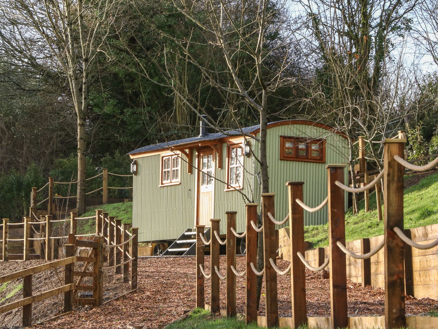 A small green corrugated metal cabin with wooden trim in a fenced outdoor area with trees at Little Silver Leaf in High Bickington