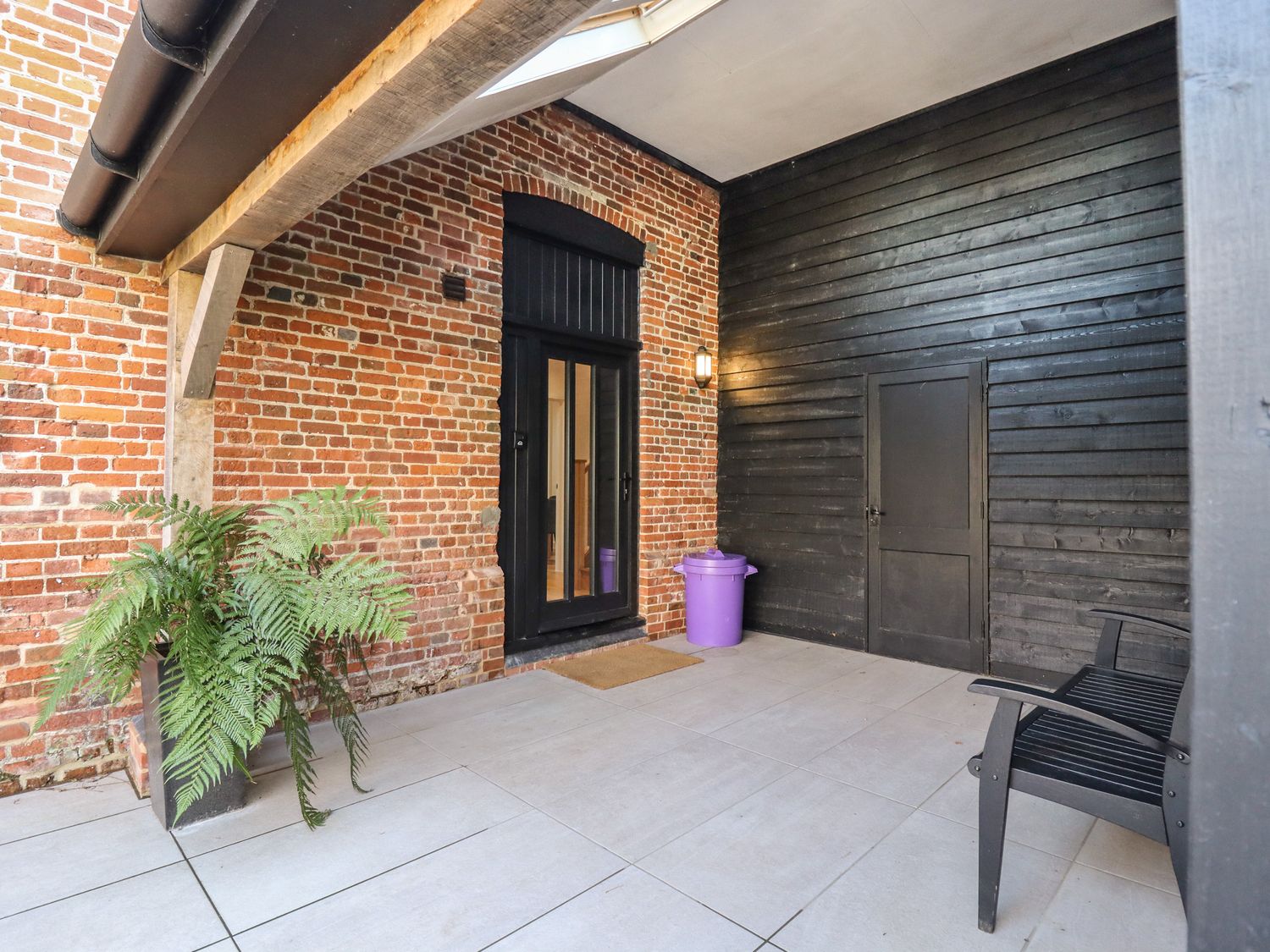 A covered outdoor patio area with brick and black wooden walls a black door a black chair a potted plant and a purple trash bin at Grange Barn in Gedding near Felsham