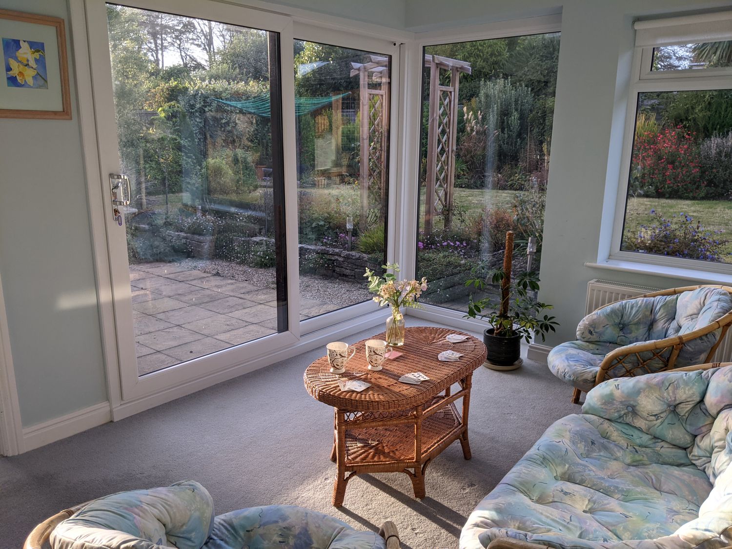 A sunroom with wicker furniture and a small coffee table with cups and flowers overlooking a garden at Connaught Gardens in Weymouth