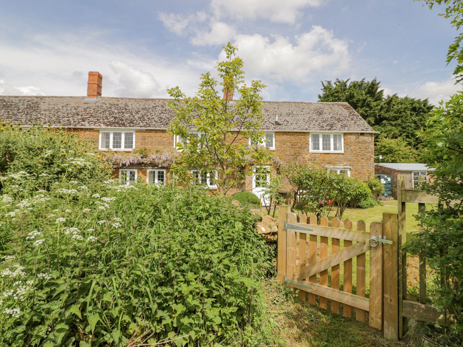 A garden with plants and a wooden gate in front of a stone house at Priors Mead in Claydon near Cropredy