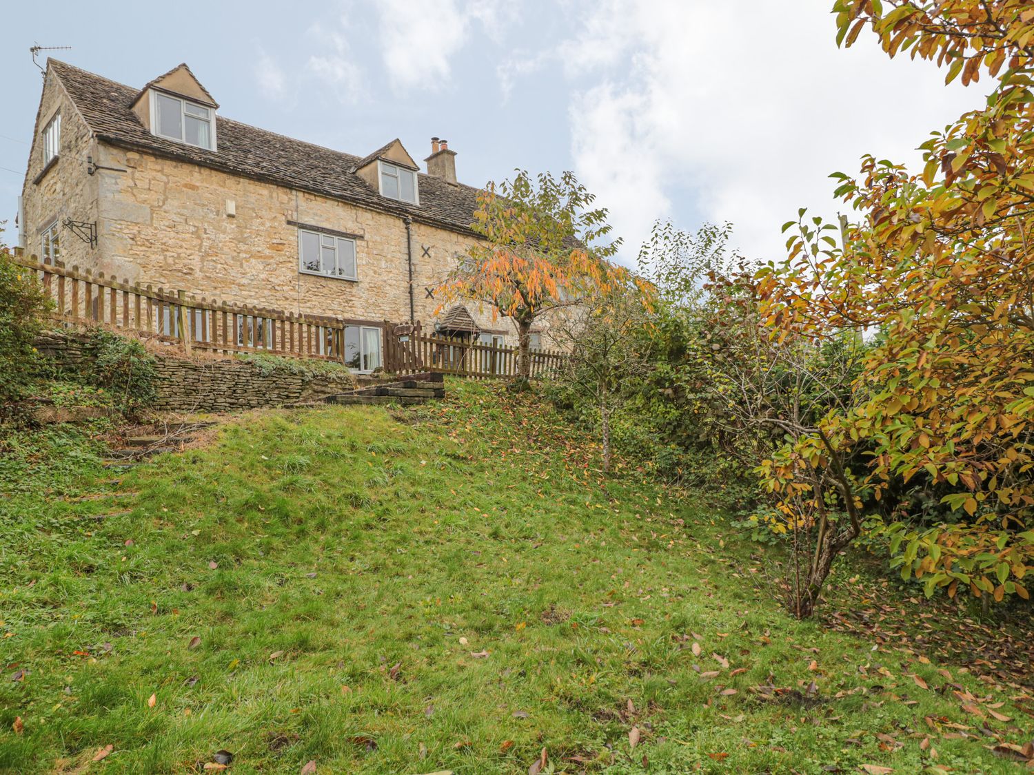 A stone house with a sloped grassy garden and autumn trees at Little End in Amberley near Stroud