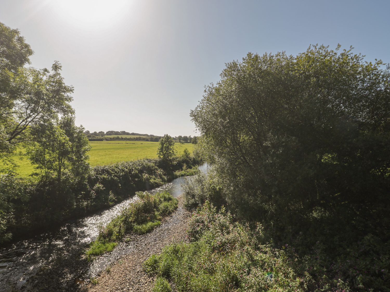 A river flowing through trees and bushes with fields and hills in the background at The Barn in Honiton