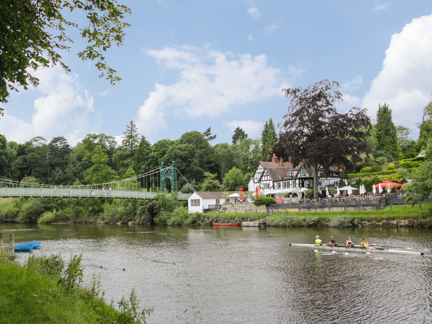 A river with rowers a suspension bridge and a building with outdoor seating at Willow View in Shrewsbury