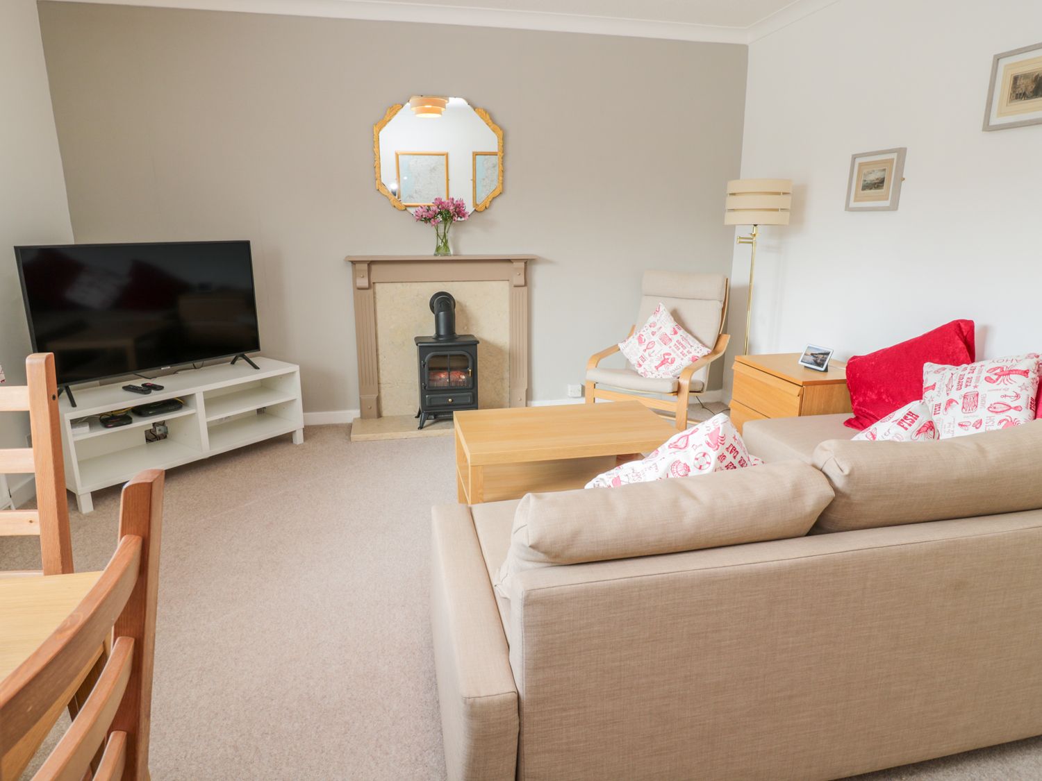 A living room with a beige sofa and armchair a wooden coffee table a television on a white stand and a fireplace with a mirror above at The Wynd Apartment in Amble