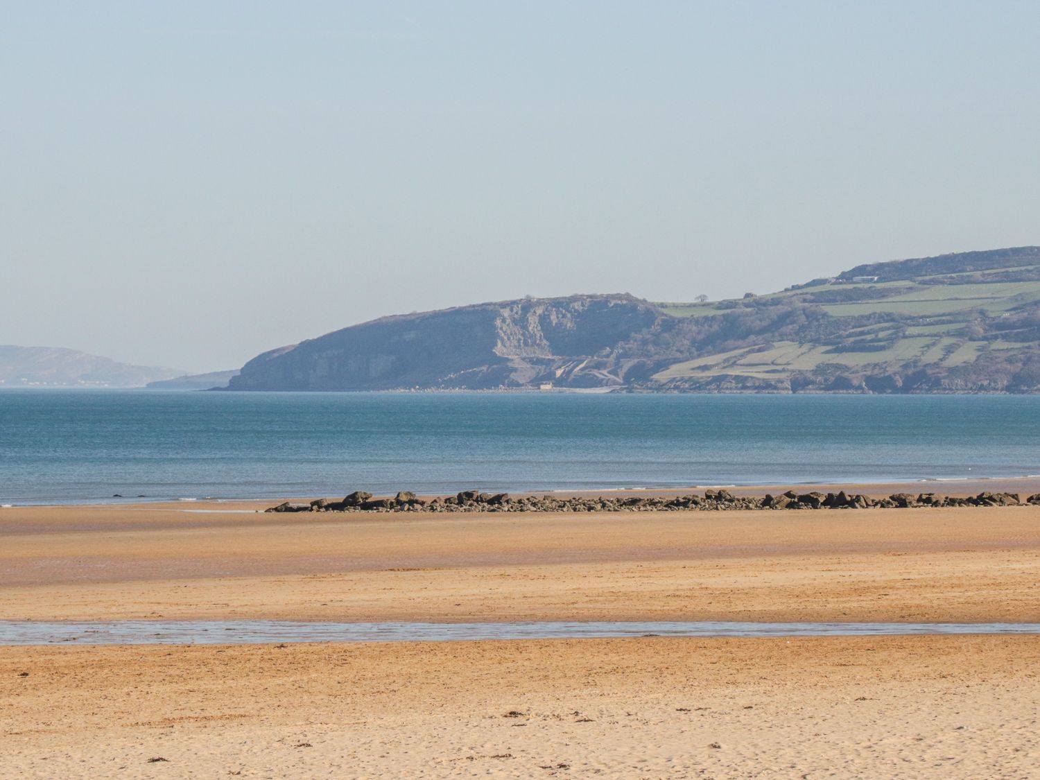 A sandy beach with a rocky barrier and ocean with hills in the background at The Penthouse in Benllech
