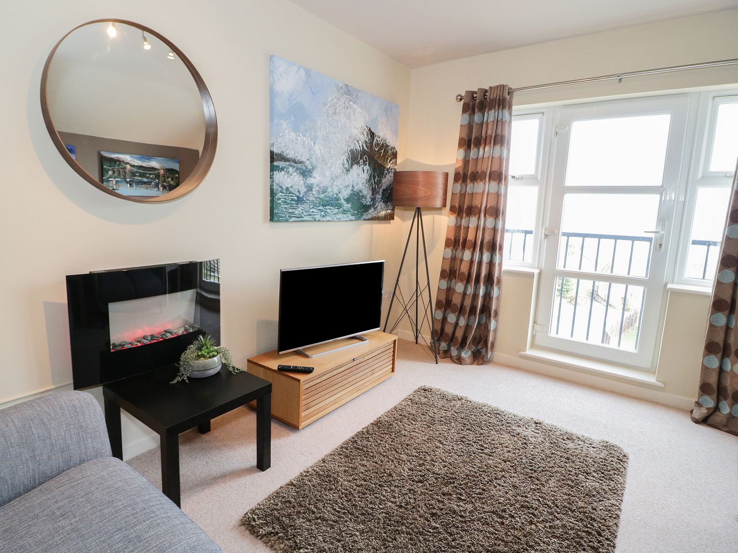 A living room with a television on a wooden stand a black side table with a plant a round wall mirror a floor lamp polka dot curtains and a rug at The Penthouse in Benllech