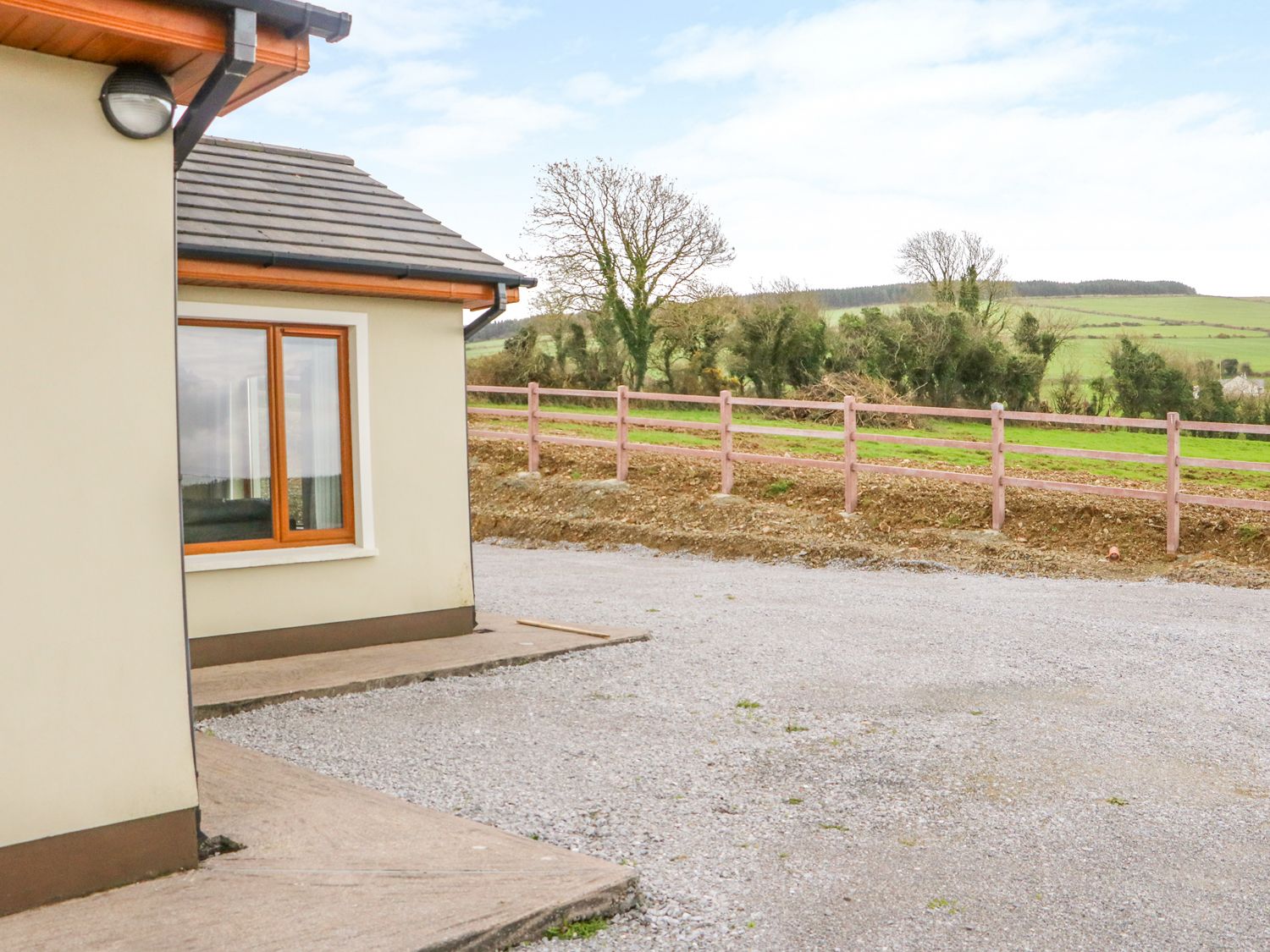 Exterior corner of a house with window facing a gravel driveway and wooden fence with fields and trees beyond at Rossanean in Farranfore near Killarney County Kerry