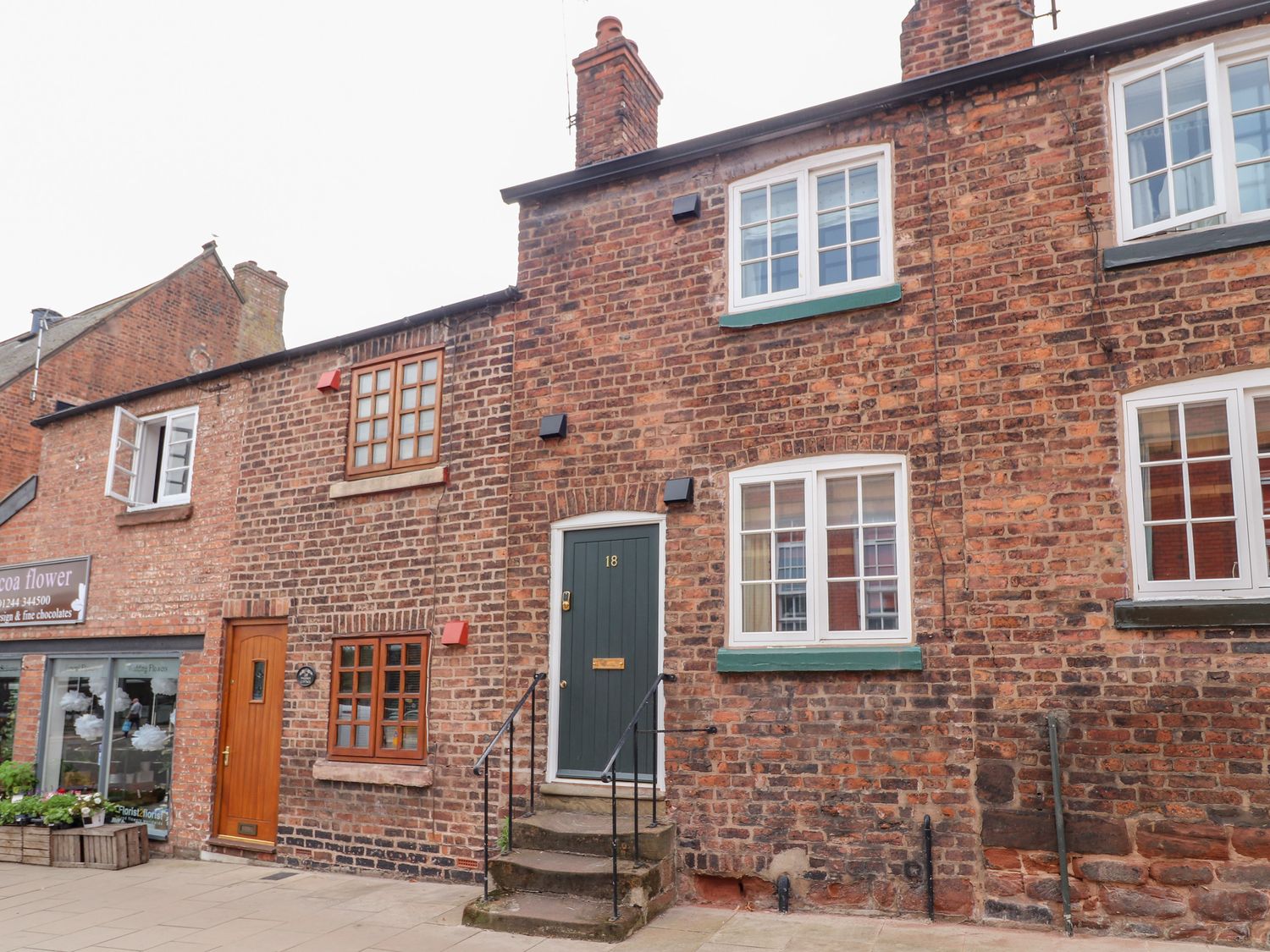 A brick building with windows and a green door numbered 18 next to a shop with flowers in the window at Tower View Cottage in Chester