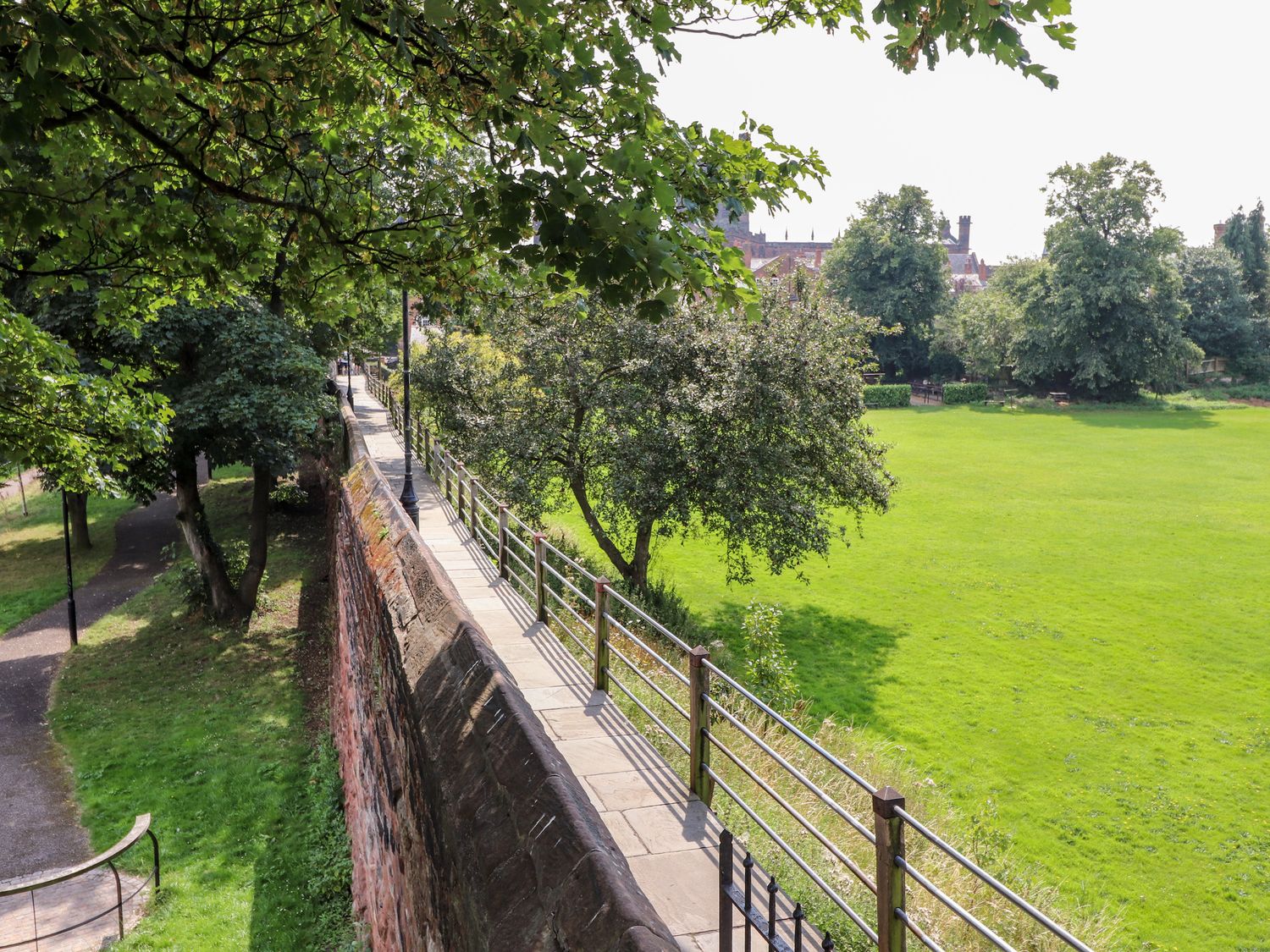 A stone walkway with metal railing along a wall overlooking a green park with trees at Tower View Cottage in Chester