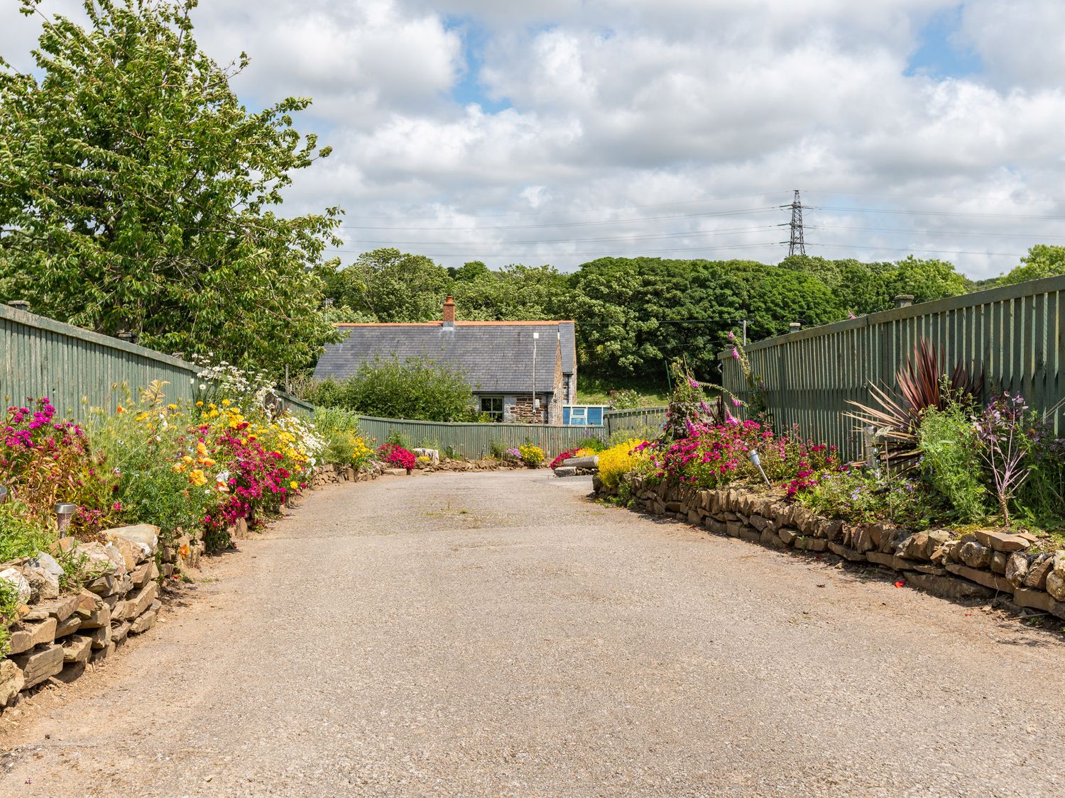 A paved pathway lined with flower beds and green fences leading to a stone building with a slate roof at Be Our Guest Cottage in Chacewater near Truro