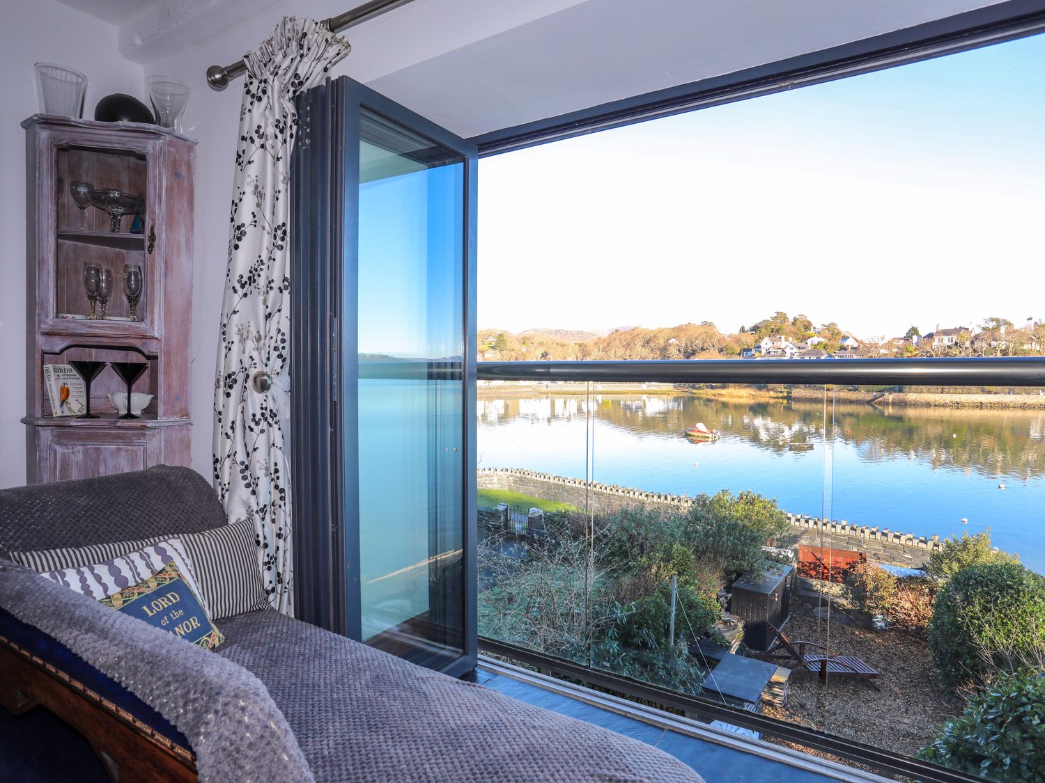 A room with an open glass door showing a view of a river and houses at St Agnes in Borth-Y-Gest