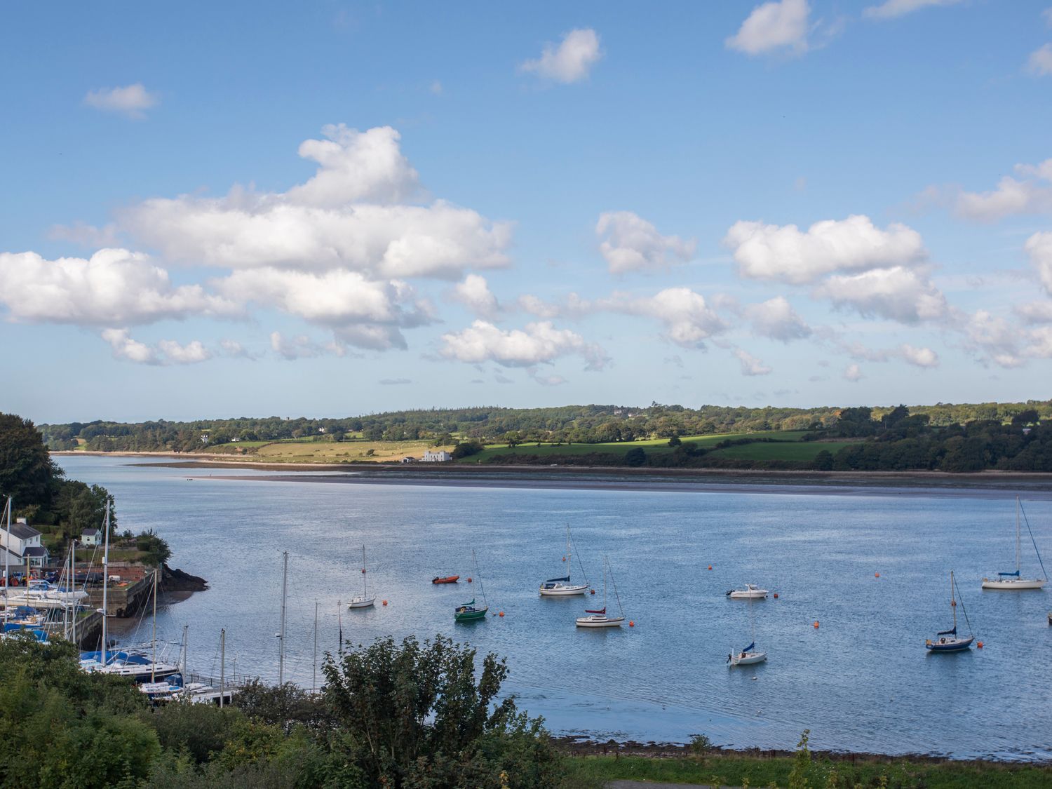 A river with boats anchored near the shore and green fields and trees on the opposite bank at Bronallt in Y Felinheli