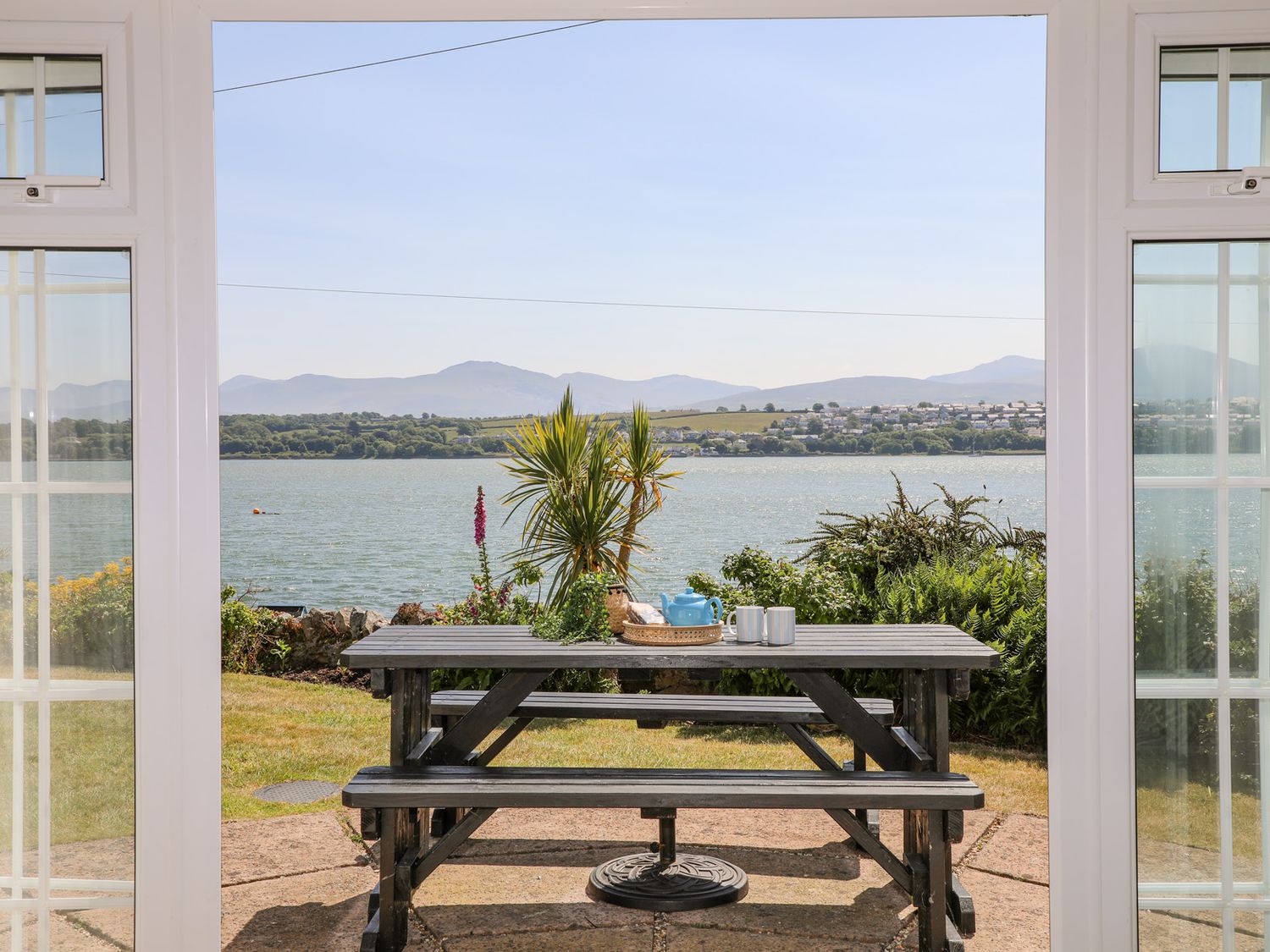 A view of a picnic table with a teapot and mugs outside overlooking water hills and houses at Mermaid Cottage in Brynsiencyn
