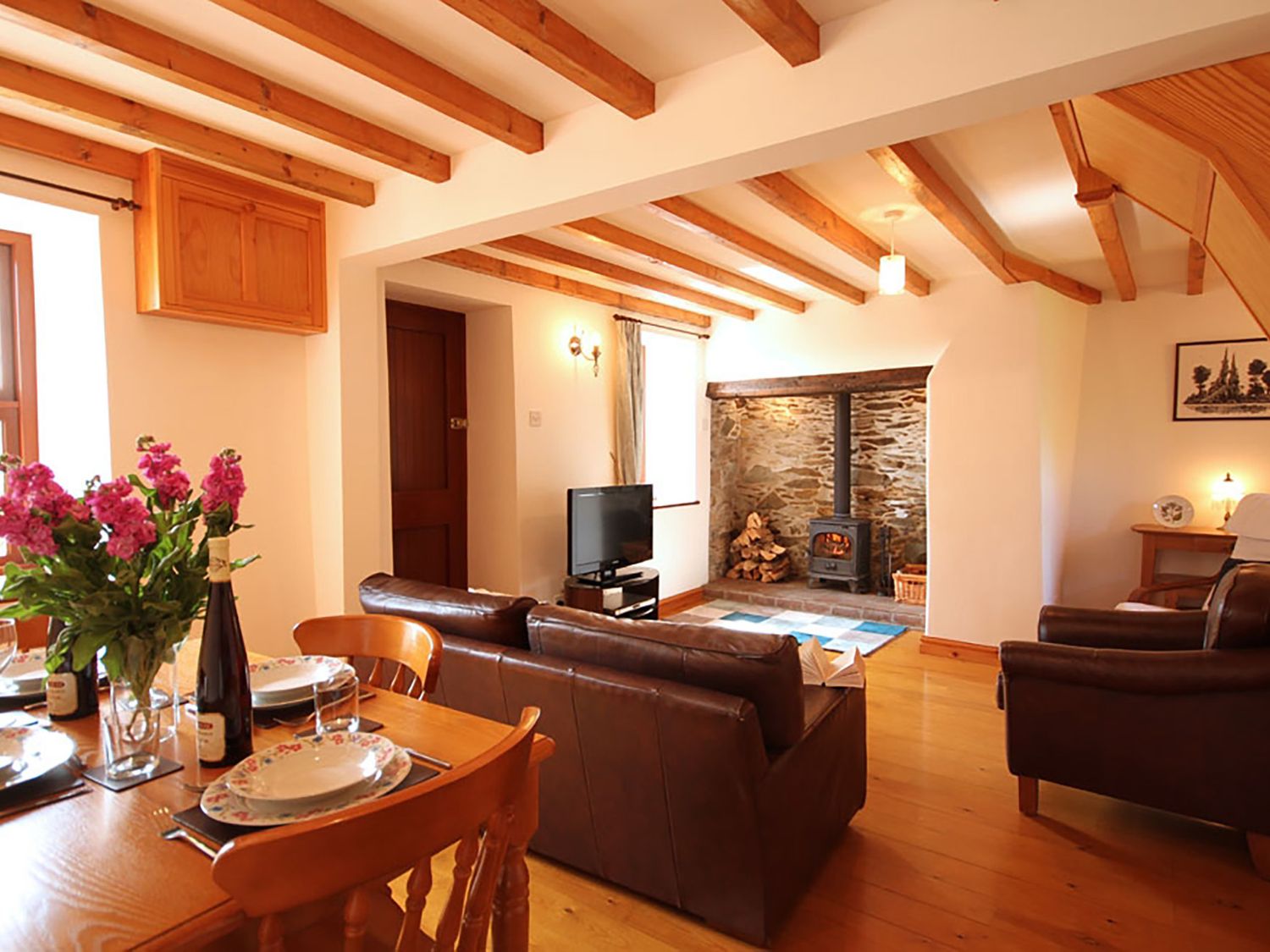 A living room with wooden ceiling beams and a stone fireplace next to a dining table with flowers at Tyn y Mynydd in Bull Bay