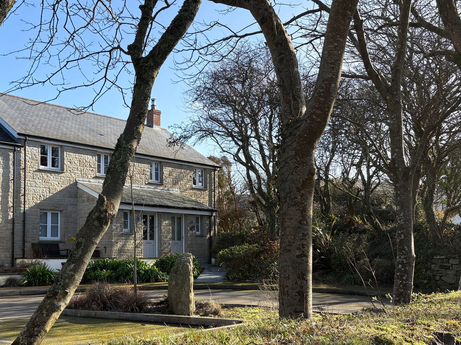 A stone house with multiple windows surrounded by trees and plants in a garden area at Tinners Rest in Pendeen