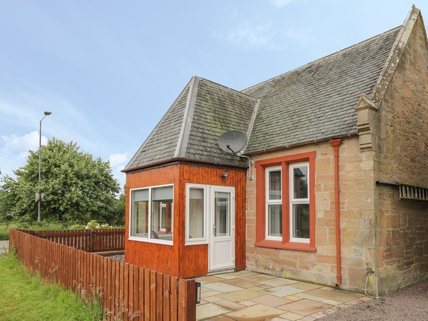 An exterior view of a stone house with a wooden enclosed porch and a fenced yard at Blantyre Cottage in Muir of Ord