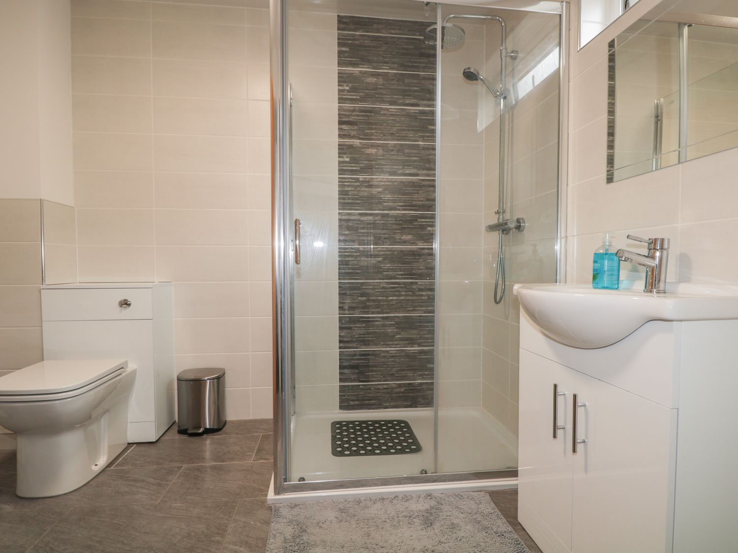 A bathroom with a toilet a trash bin a shower area with glass doors and a sink with a mirror at Blantyre Cottage in Muir of Ord