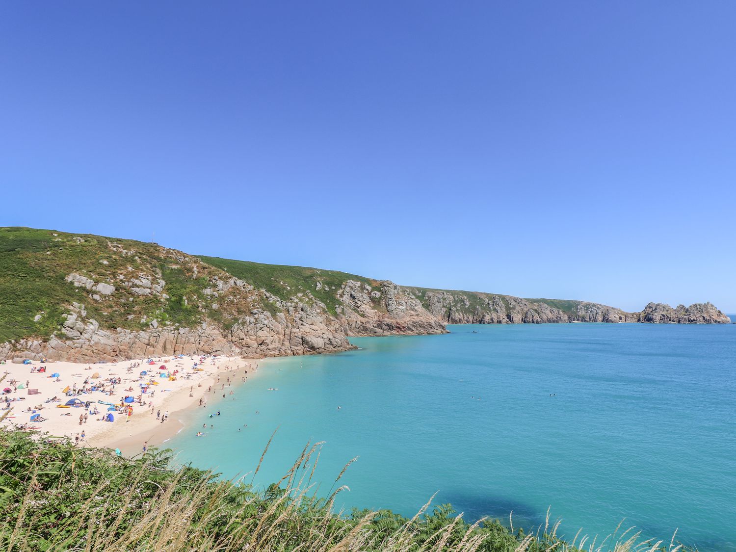 A sandy beach with people swimming and sunbathing near rocky cliffs and blue sea at The Upper Deck in Porthcurno