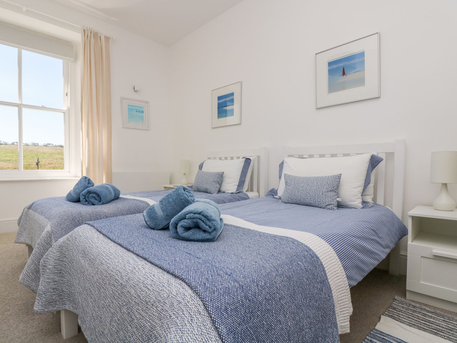 A bedroom with two single beds with blue bedding and rolled towels a window and framed pictures on the wall at The Upper Deck in Porthcurno