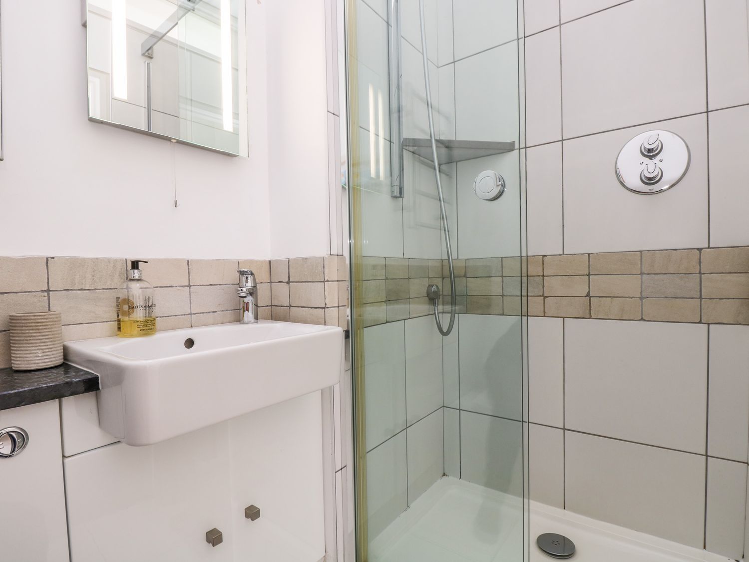 A bathroom with a sink, mirror, soap dispenser, and a glass-enclosed shower at The Upper Deck in Porthcurno