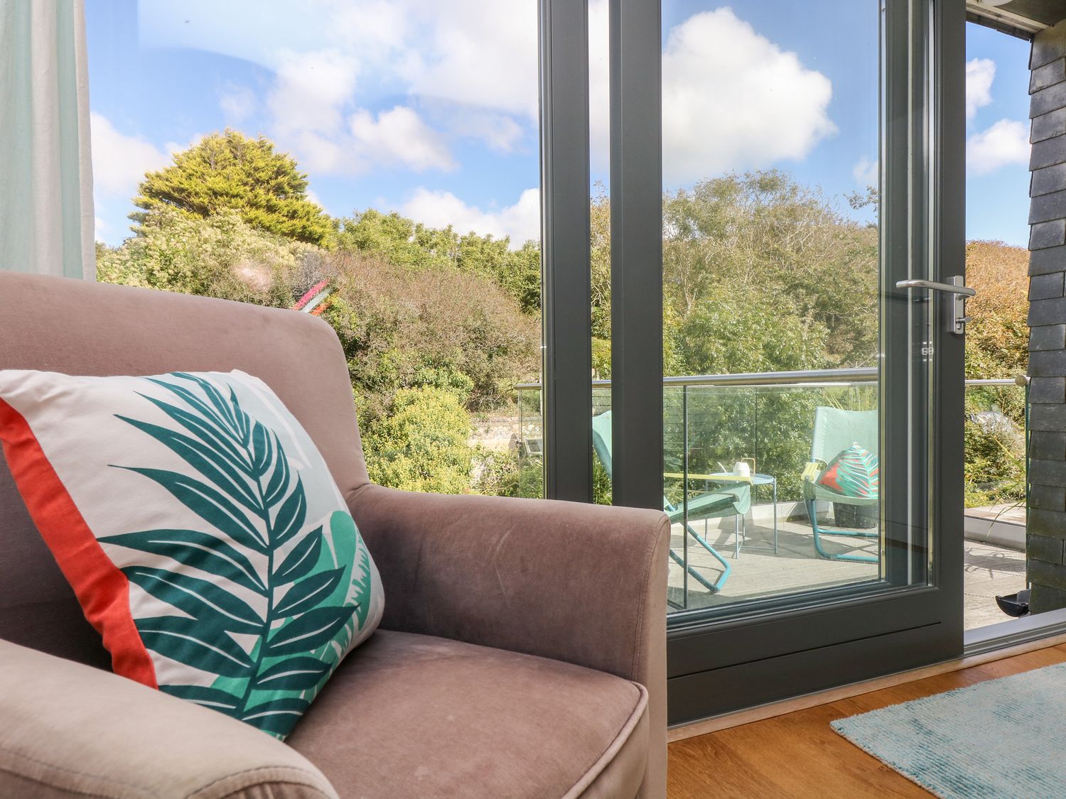 An armchair with a leaf patterned cushion next to a glass door leading to a balcony with chairs at The Upper Deck in Porthcurno