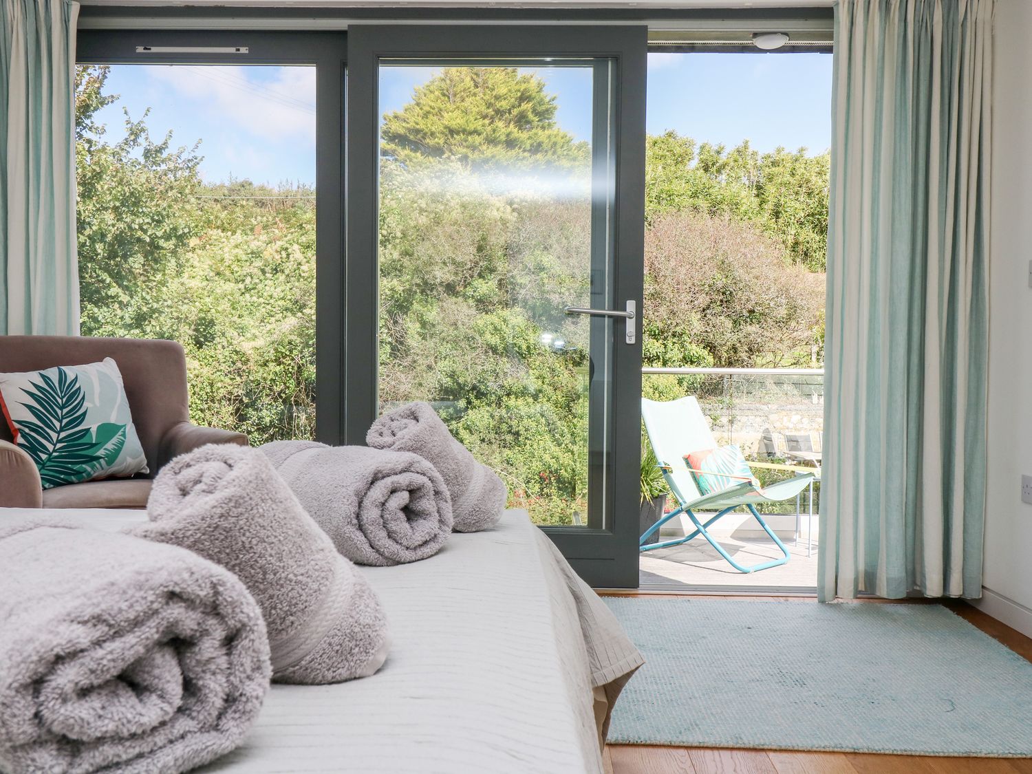 A bedroom with rolled towels on the bed and a chair near glass doors leading to a balcony at The Upper Deck in Porthcurno