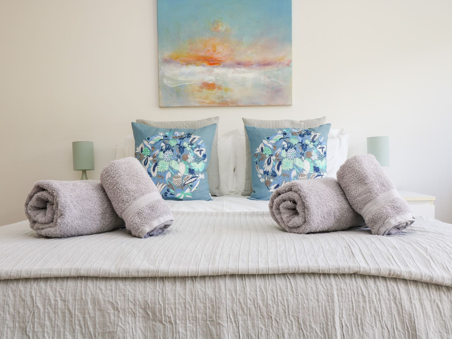 A bed with rolled towels and floral pillows under a painting at The Upper Deck in Porthcurno