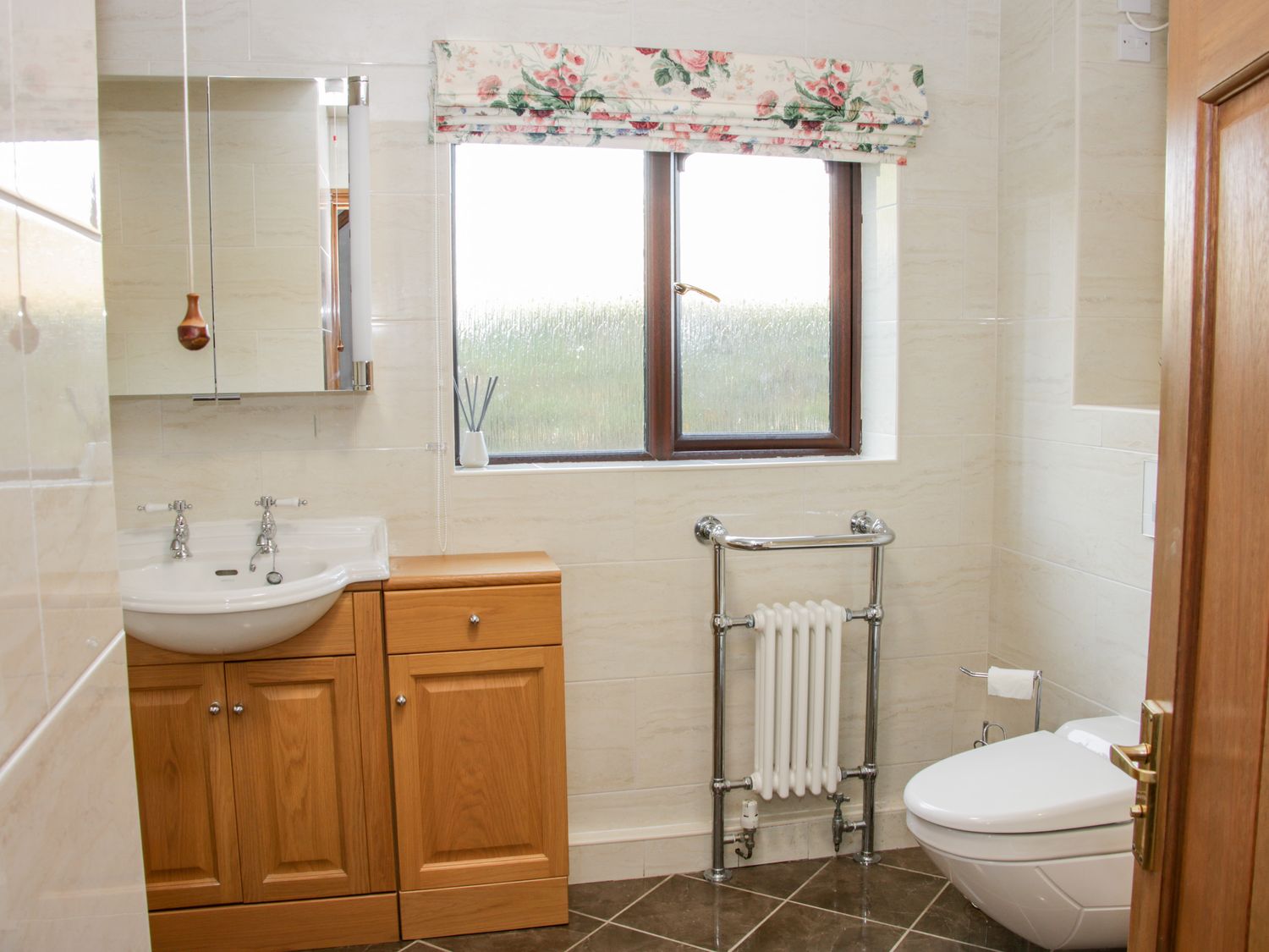 A bathroom with a sink and wooden cabinet a window with floral blinds a towel radiator and a toilet at Court Farm in Neen Sollars near Cleobury Mortimer