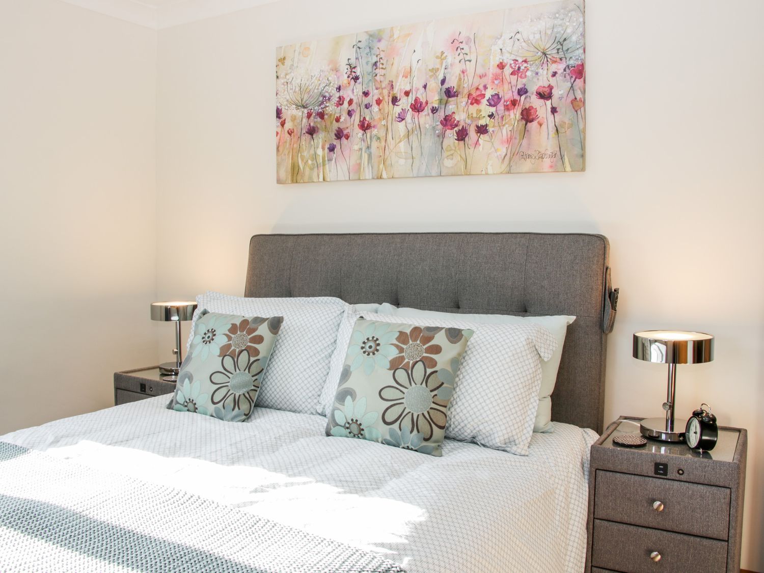 A bedroom with a grey upholstered bed with patterned pillows and two nightstands with lamps and an alarm clock at Court Farm in Neen Sollars near Cleobury Mortimer