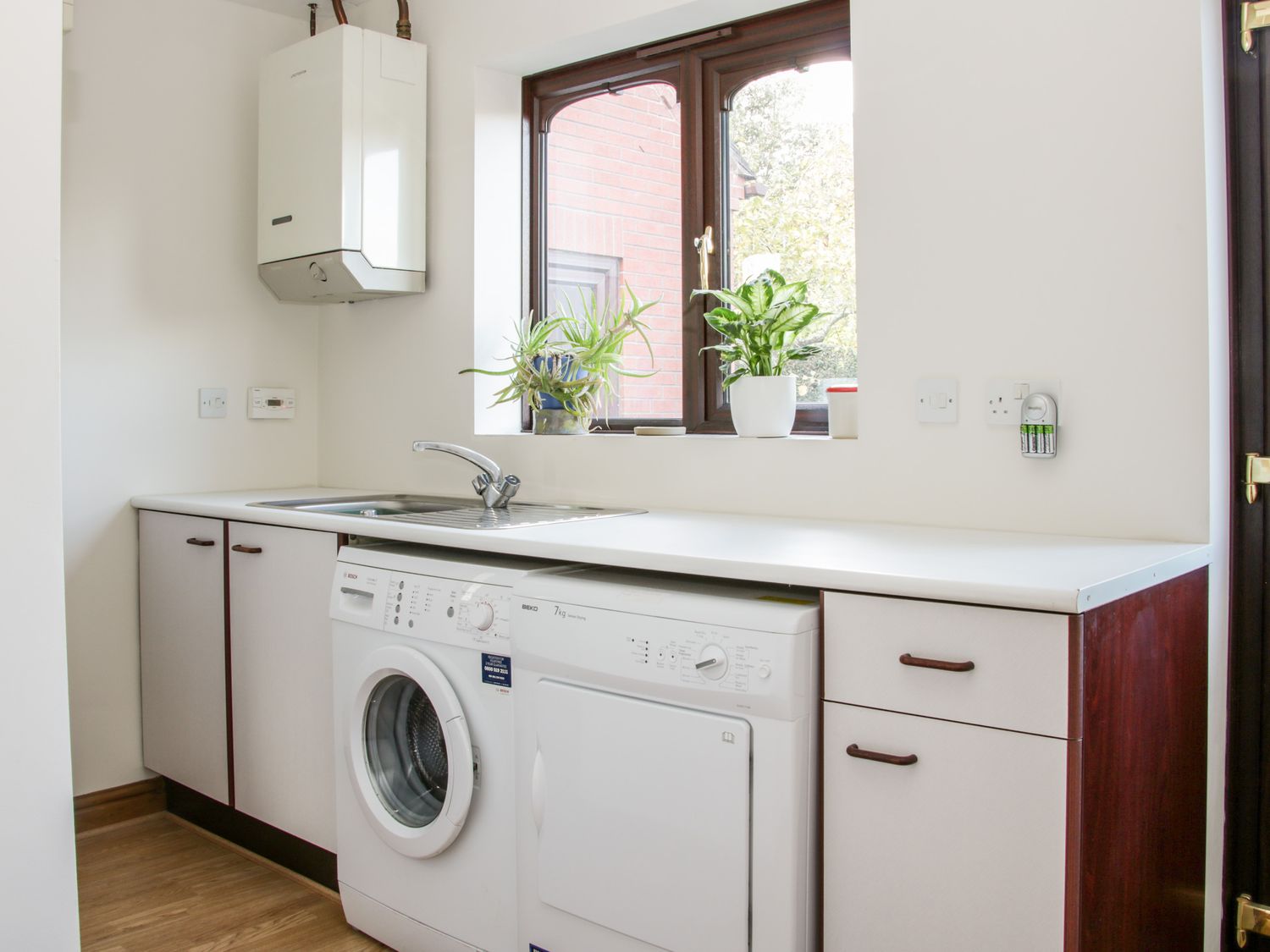A utility room with a washing machine, dryer, sink, cabinets, and a window with plants on the sill at Court Farm in Neen Sollars near Cleobury Mortimer
