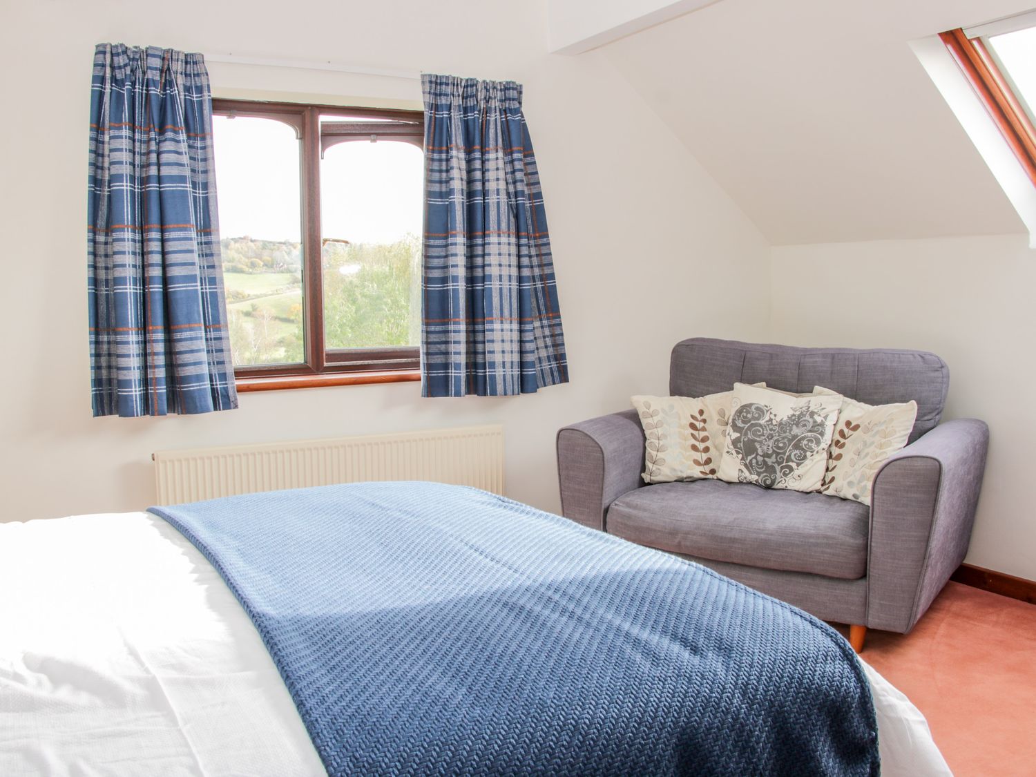 A bedroom with a bed covered by a blue blanket a gray armchair with cushions and a window with blue plaid curtains at Court Farm in Neen Sollars near Cleobury Mortimer