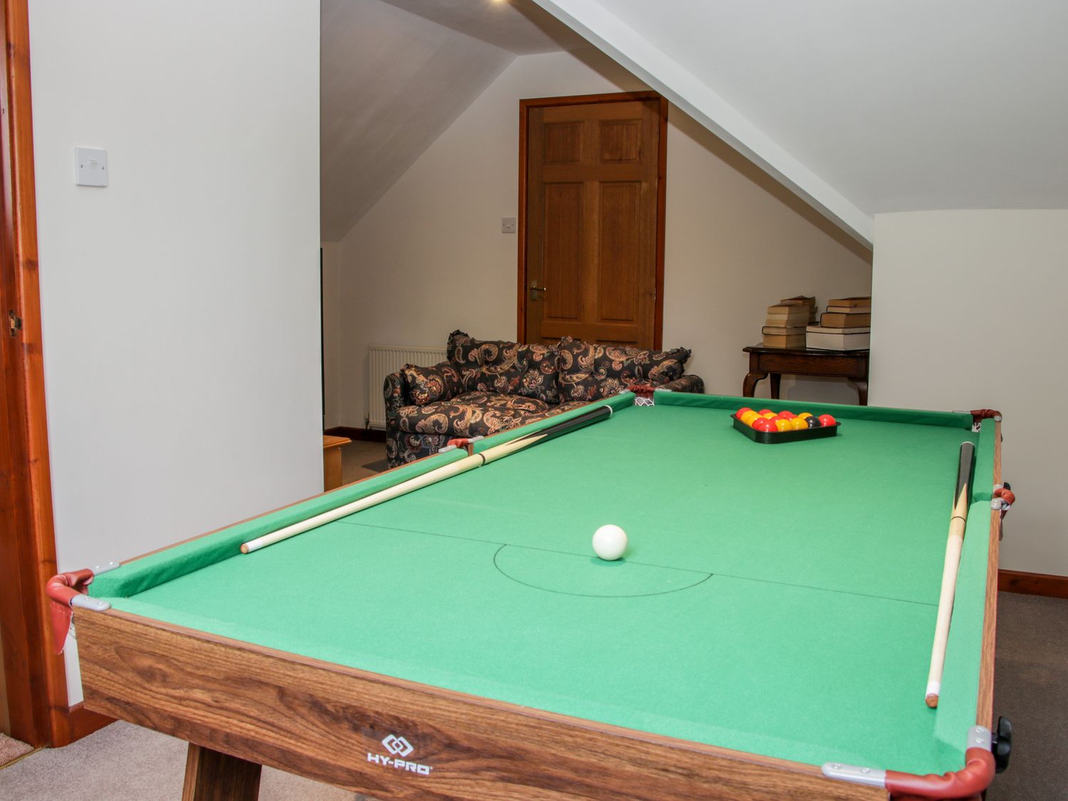 A room with a green pool table two pool cues a patterned sofa a wooden door and a table with stacked books at Court Farm in Neen Sollars near Cleobury Mortimer