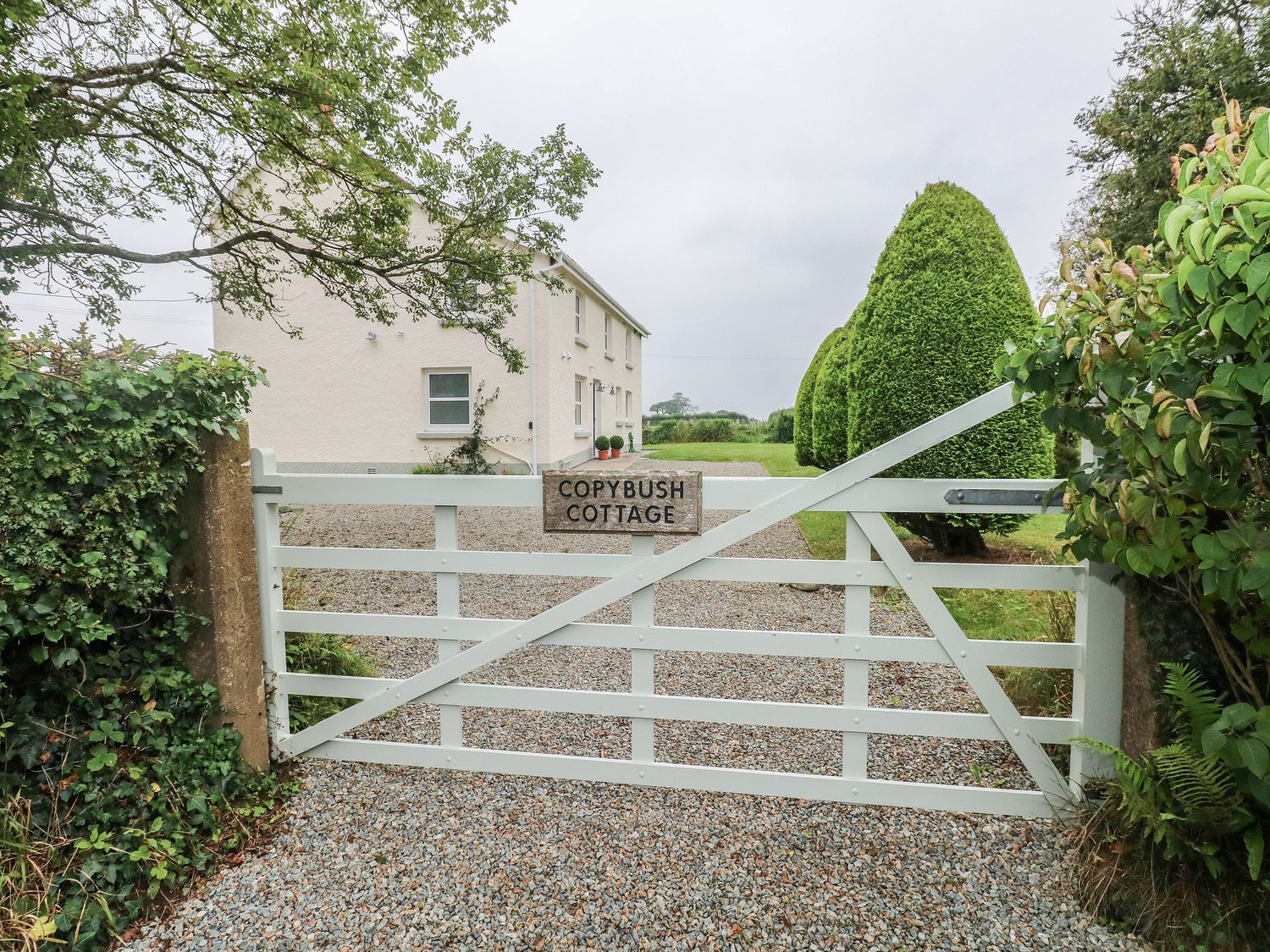 A white wooden gate with a sign reading Copybush Cottage leading to a gravel driveway and a cream house surrounded by trimmed bushes at Copybush Cottage in Lawrenny