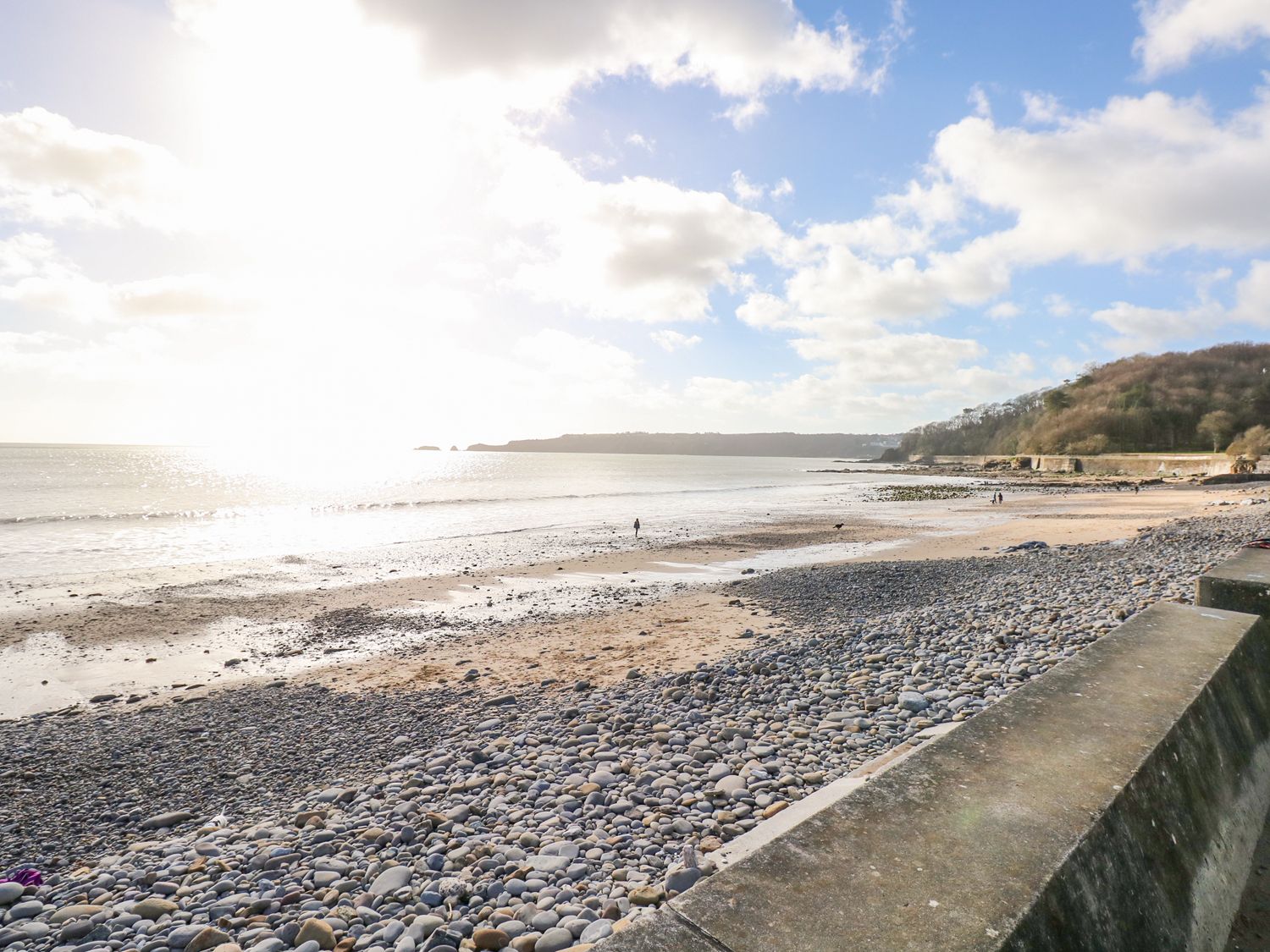 A pebble and sandy beach with a few people walking and a wooded hill near the shore at Serendipity - Heritage Park Stepaside near Kilgetty