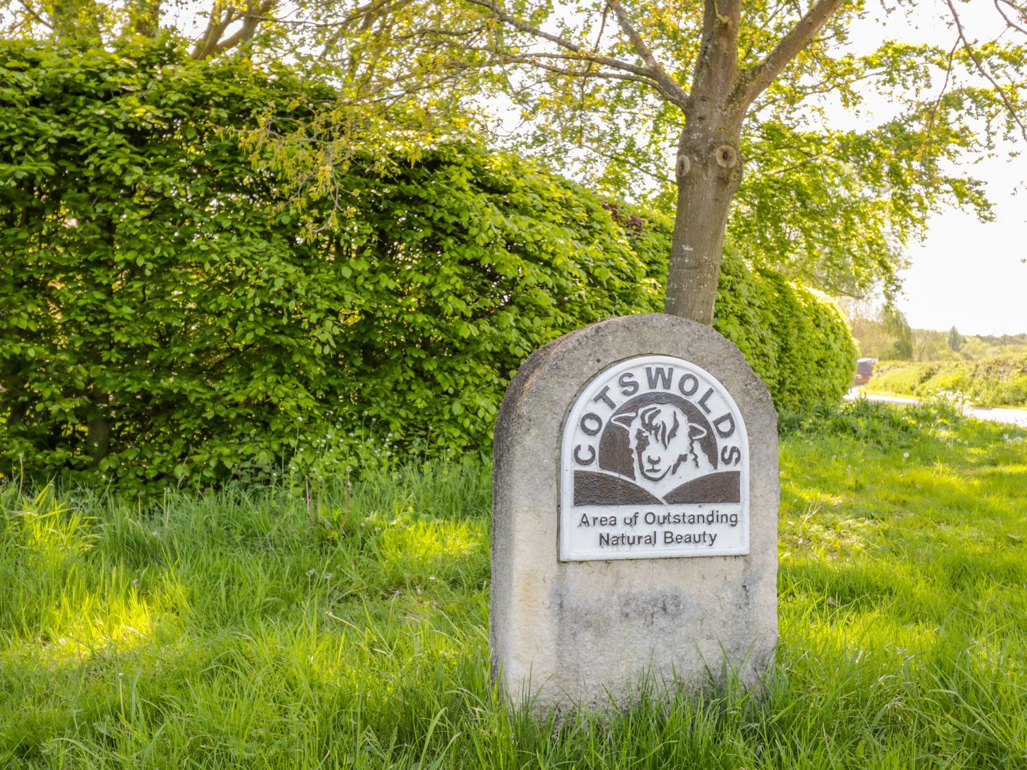 A stone sign with a sheep emblem reading Cotswolds Area of Outstanding Natural Beauty near a tree and hedge in the countryside