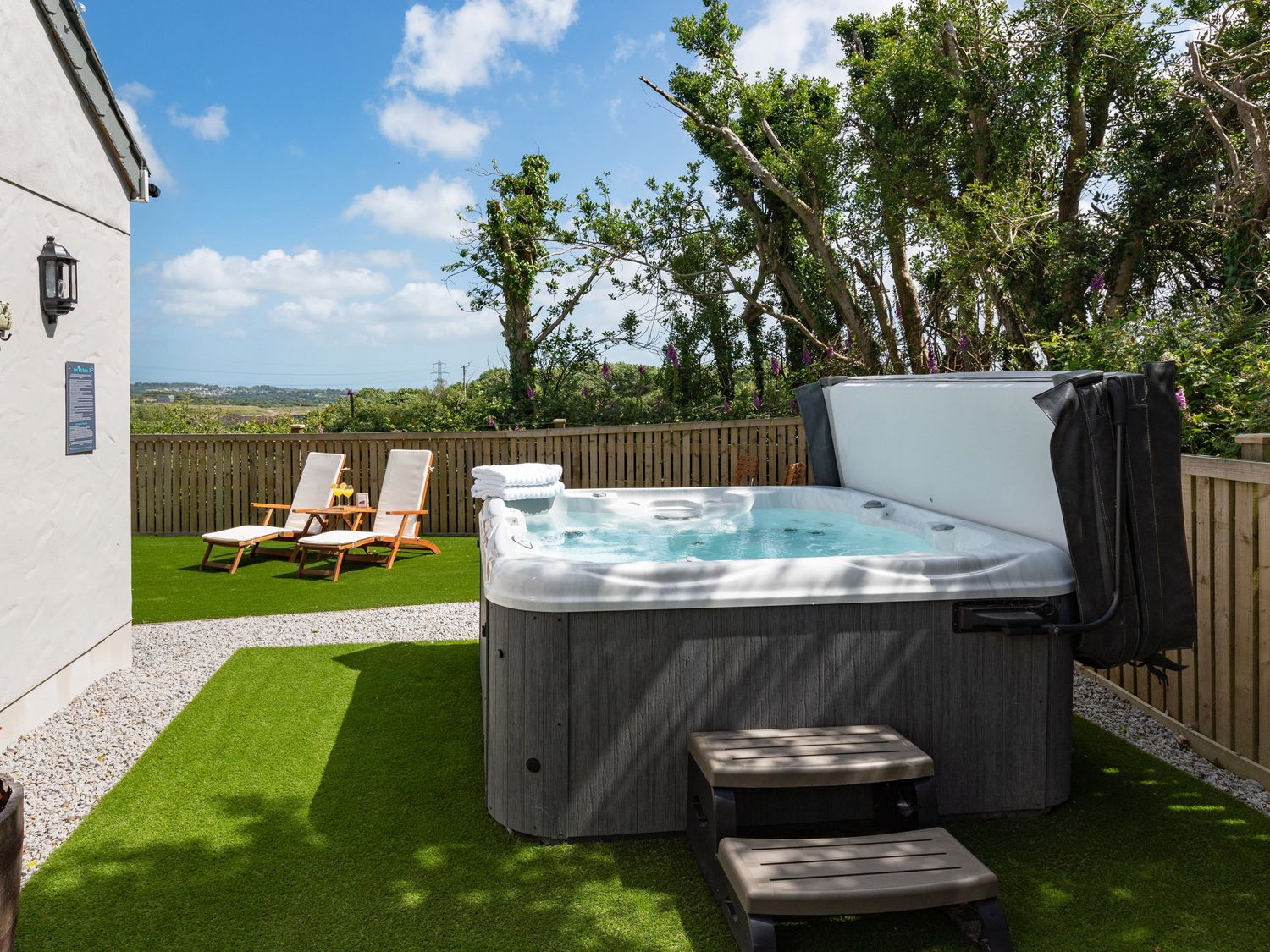 An outdoor area with a hot tub, two lounge chairs, and a fence with trees behind at Bear Cottage in Chacewater near Truro