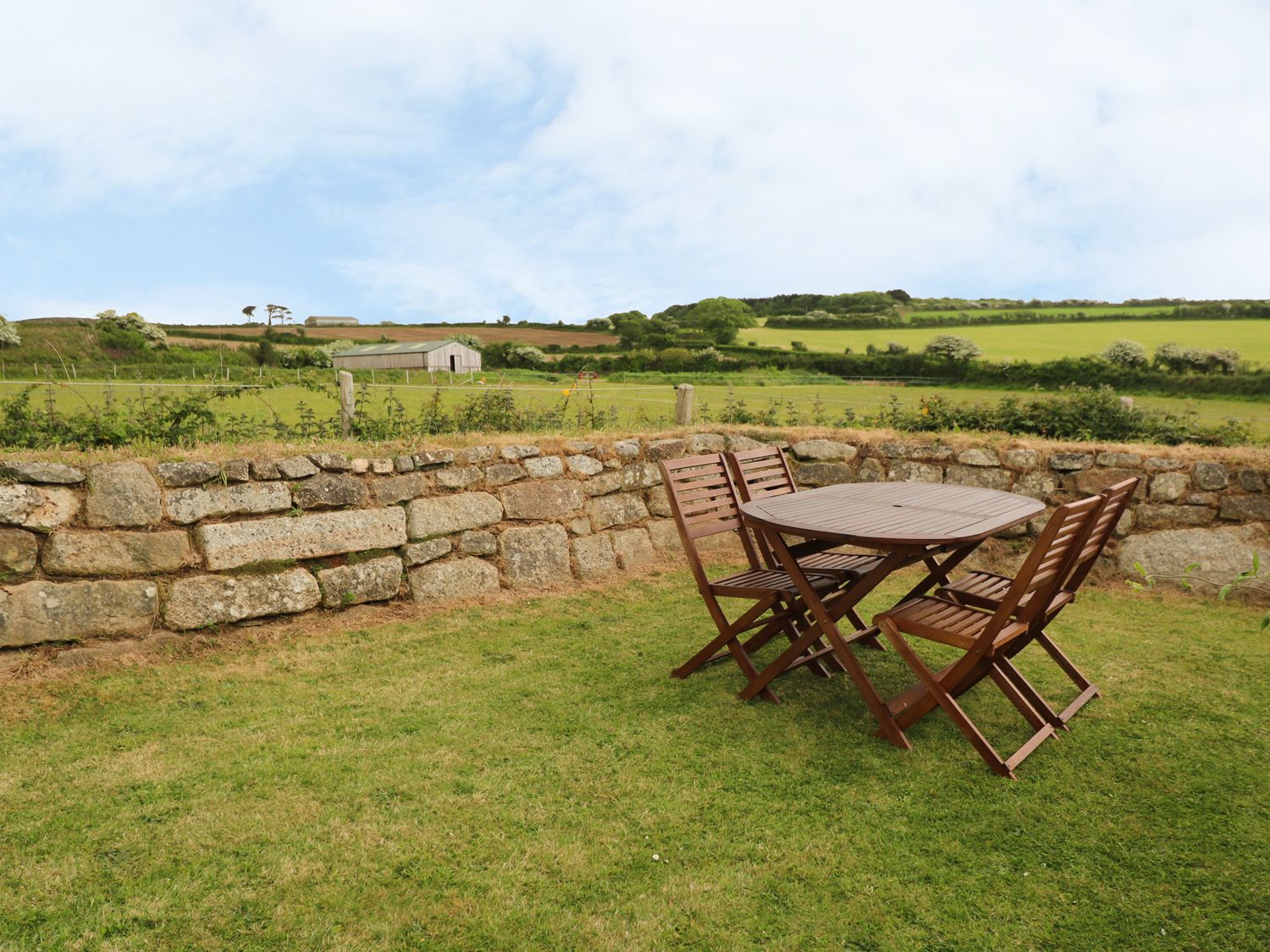 A wooden table with four chairs on grass beside a stone wall with fields and a barn in the background at Old Chapel Cottage Trescowe near Praa Sands