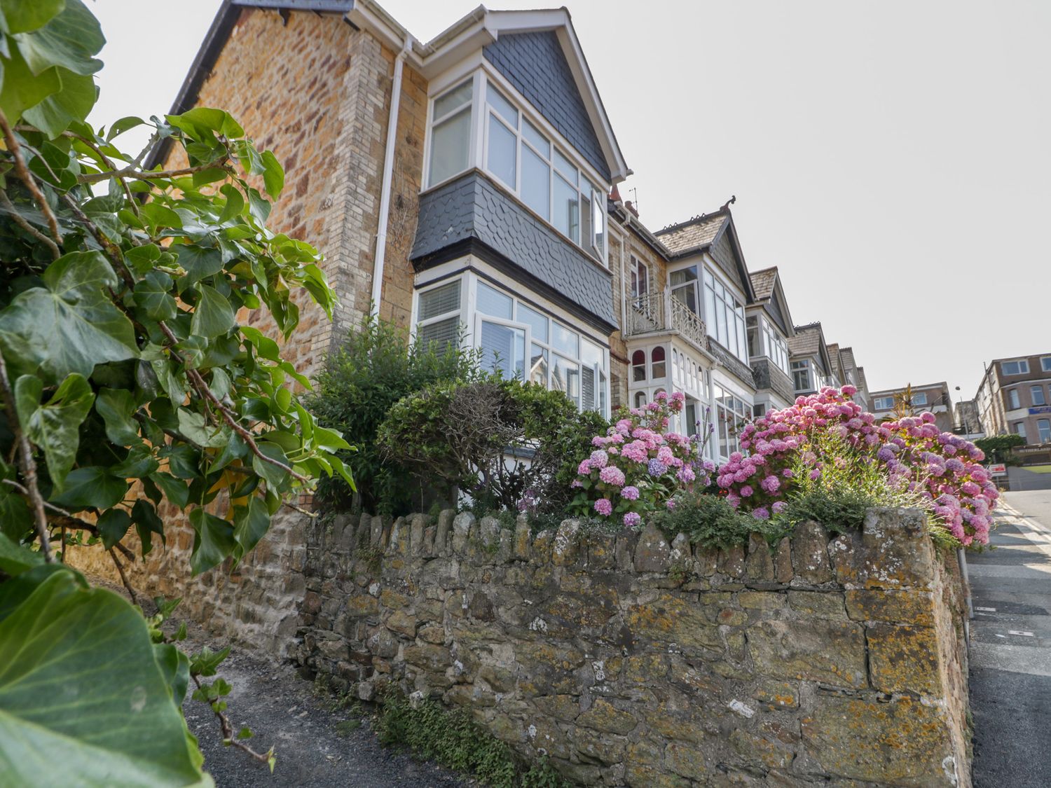 Stone houses with bay windows along a stone wall with pink flowers on a street at Seascape in Newquay