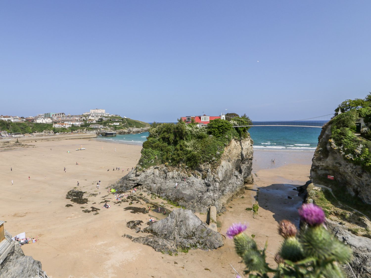 A sandy beach with people and rocky cliffs connected by a narrow bridge over the water at Seascape in Newquay