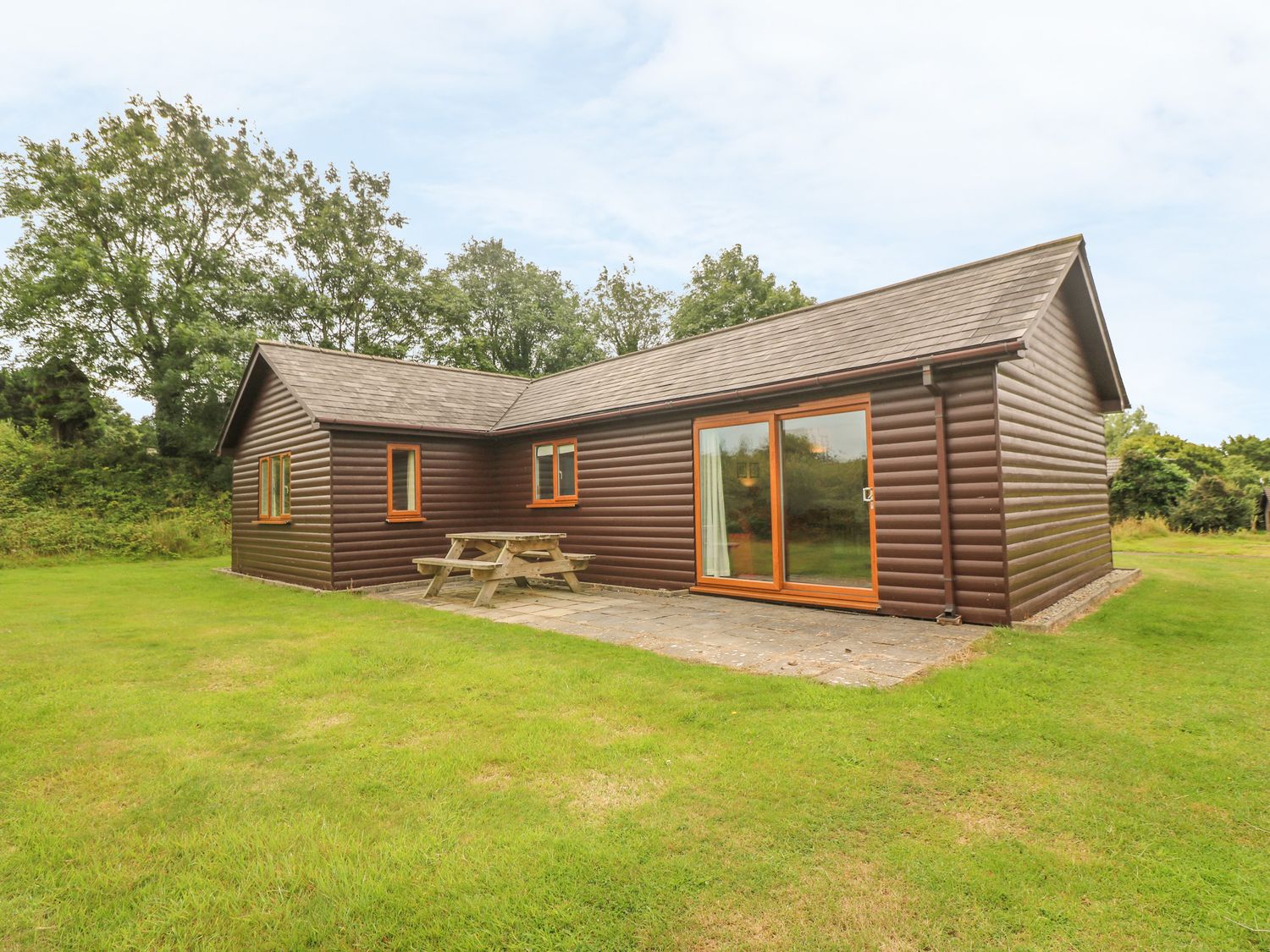 An exterior view of a wooden cabin with windows sliding door and a picnic table on a paved area surrounded by grass at 64 Waterside in Lanivet
