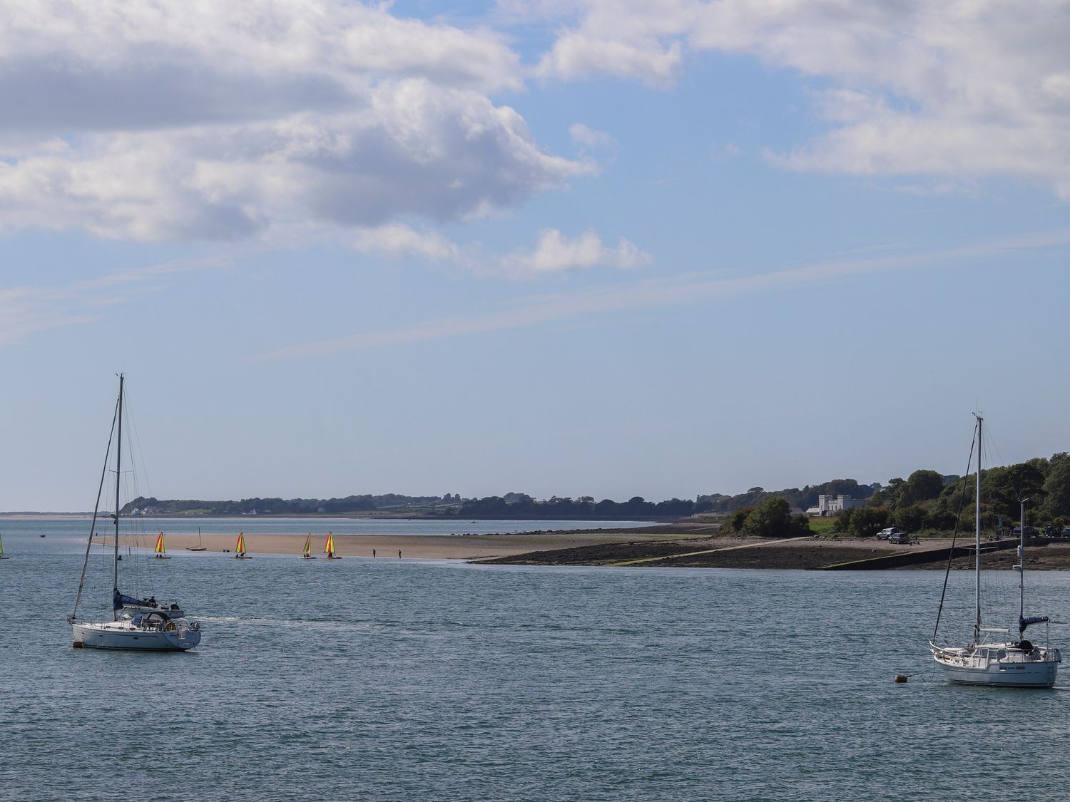 A body of water with two sailboats and small sailboats with yellow and red sails near a sandy shore at Delfryd in Y Felinheli