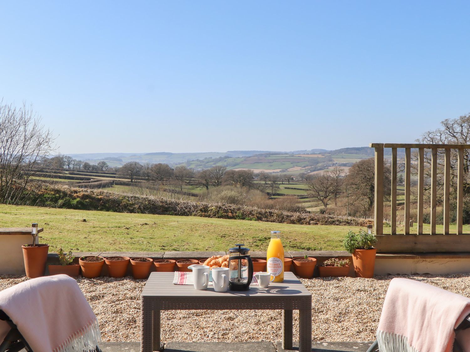 An outdoor patio with a table set with mugs a French press orange juice and croissants overlooking fields and hills at The Shippon at Wyke Farm in Lyme Regis