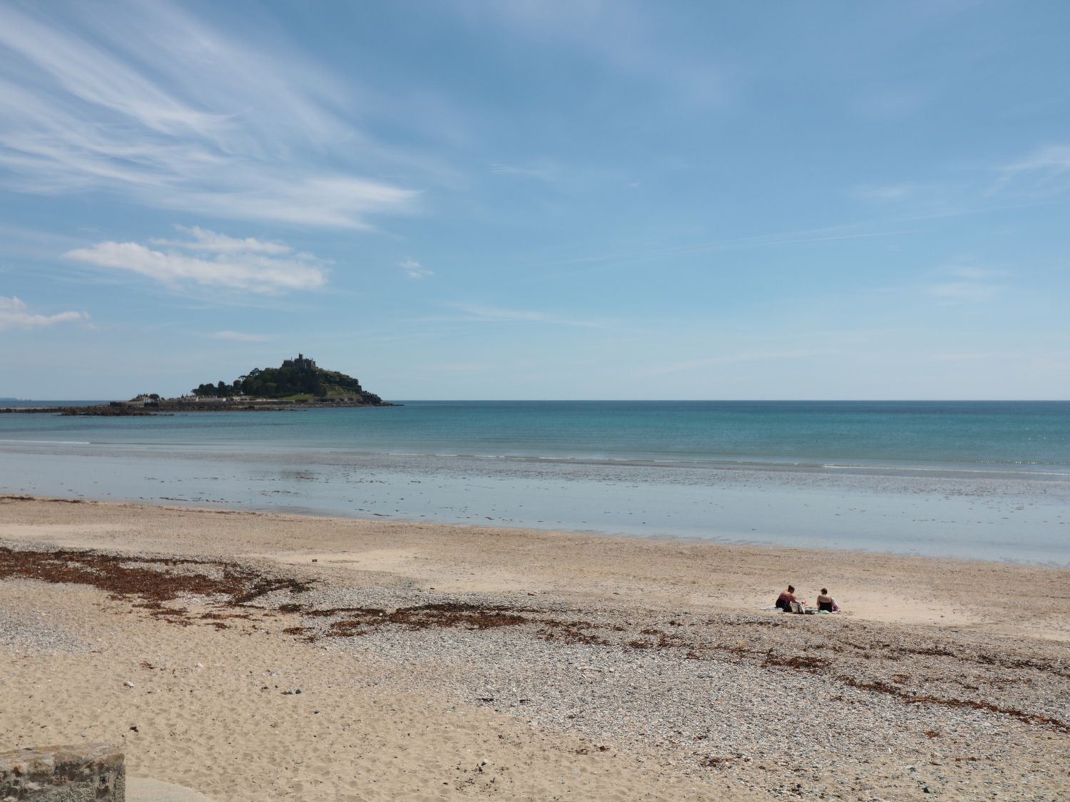 A sandy beach with two people sitting near the shore and an island with buildings in the background at Roskennals Mill in Penzance