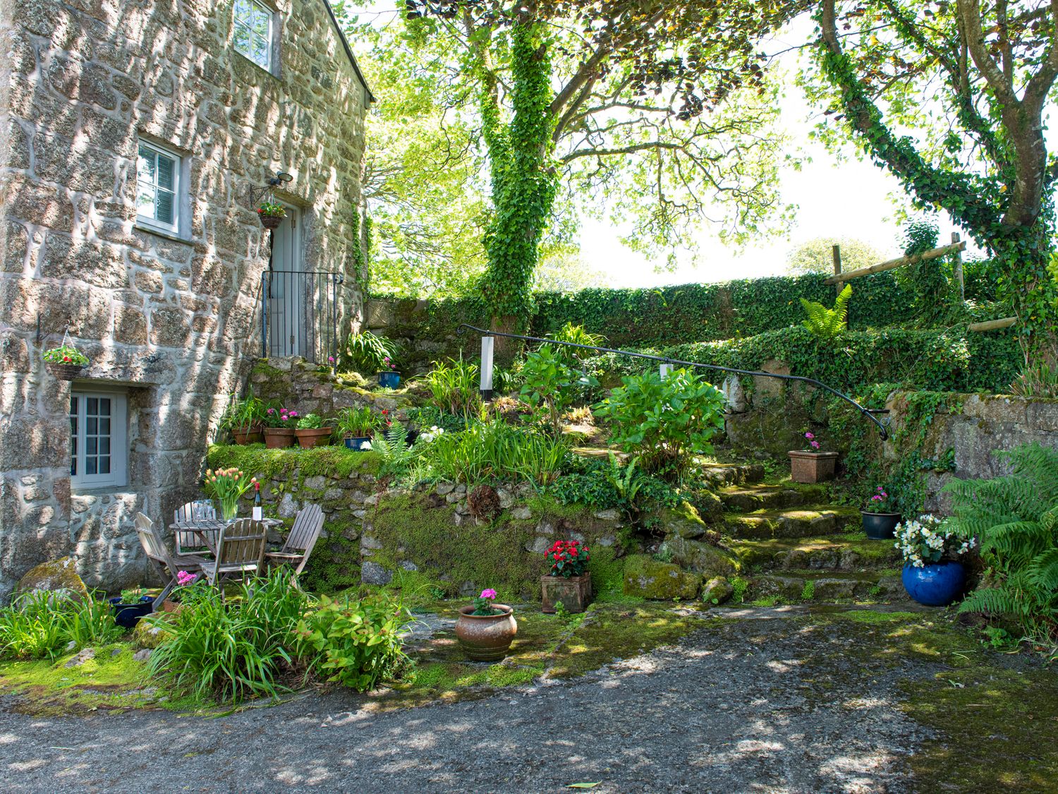 An outdoor stone patio area with wooden chairs and table next to a stone building with potted plants on steps and greenery around at Roskennals Mill in Penzance