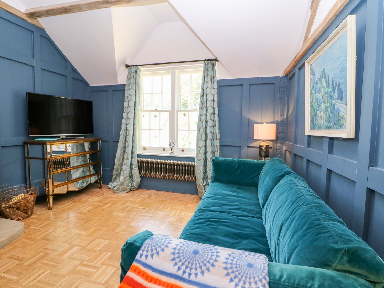 A room with blue paneled walls a teal sofa a window with curtains a television on a mirrored cabinet and a lamp at The Hemmel in Faringdon