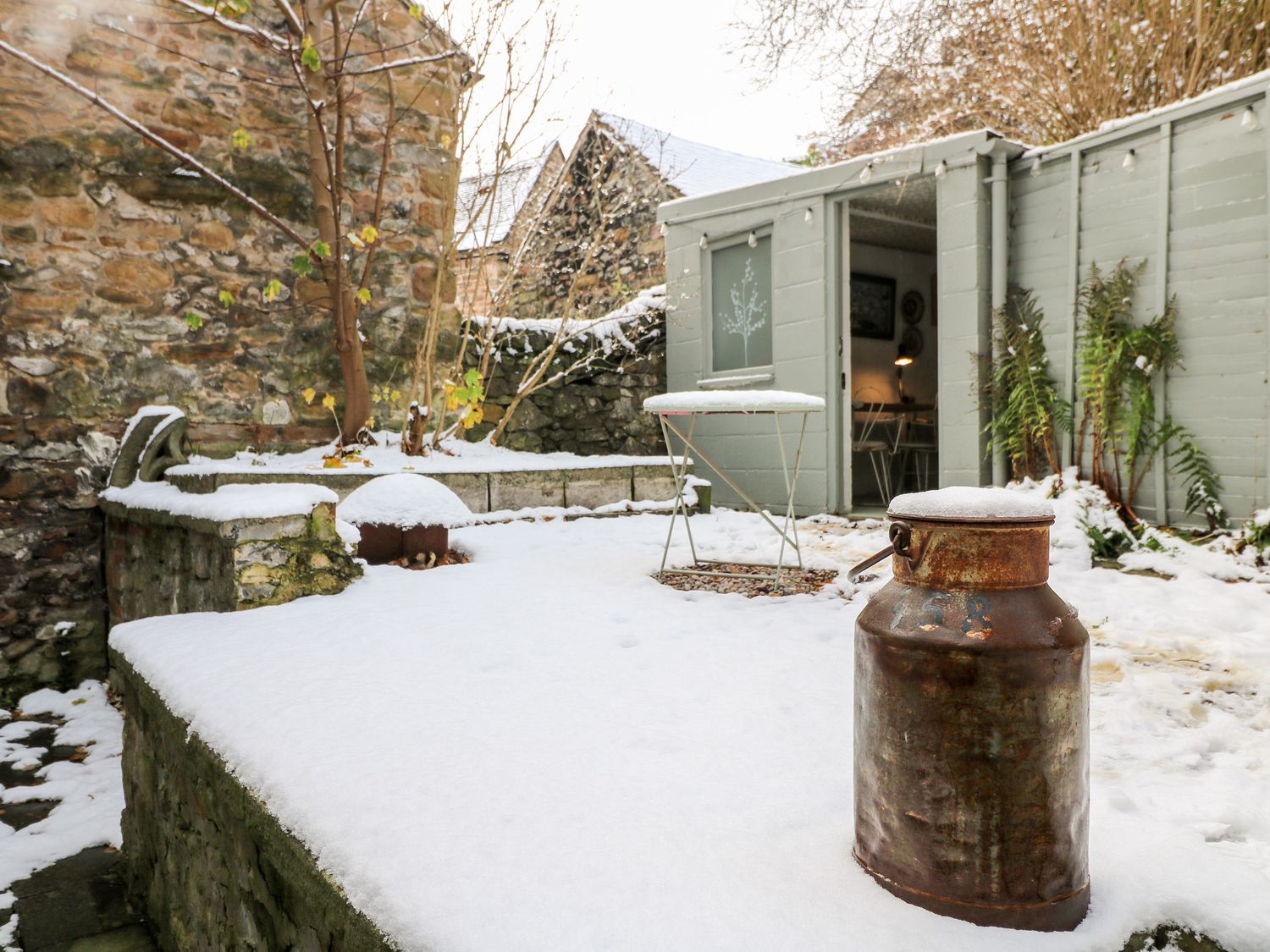 A snow-covered outdoor patio with a rusty milk can a small round table and stone walls at Church House in Wirksworth