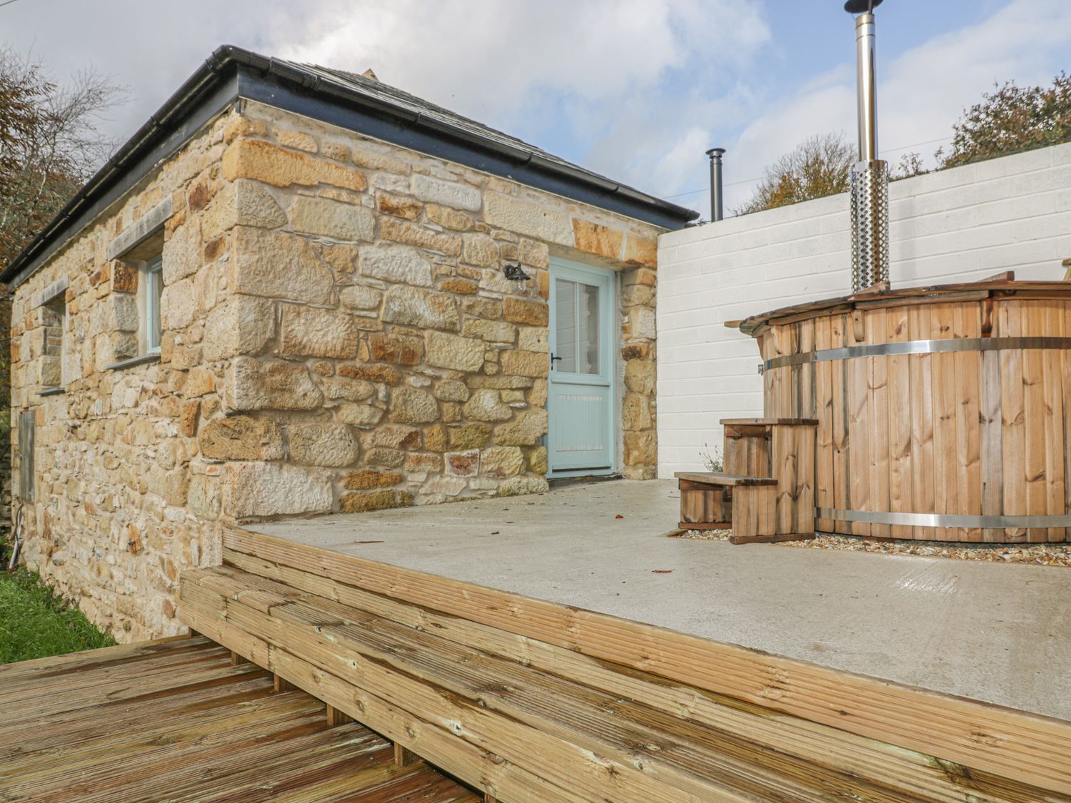 A stone building with a blue door next to a wooden hot tub on a concrete and wooden deck at The Bull Pen in Coombe near Grampound