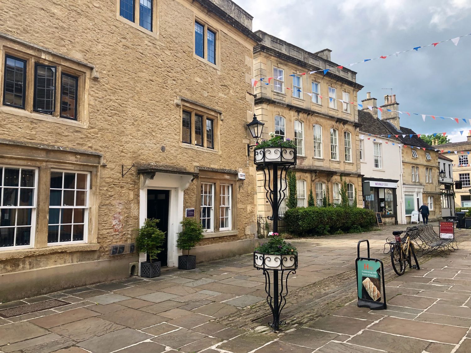 A street with stone buildings and windows with a bike parked near a lamppost with flowers at 1A Erneston Crescent in Corsham