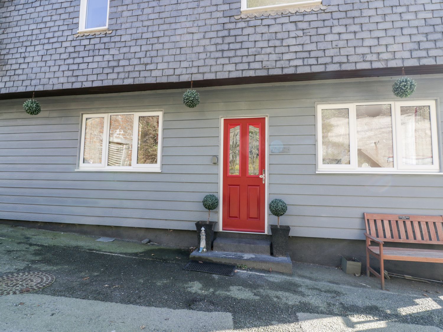 Exterior of a building with a red door two windows four hanging spherical plants and a wooden bench at Casa Del Mar in Mevagissey