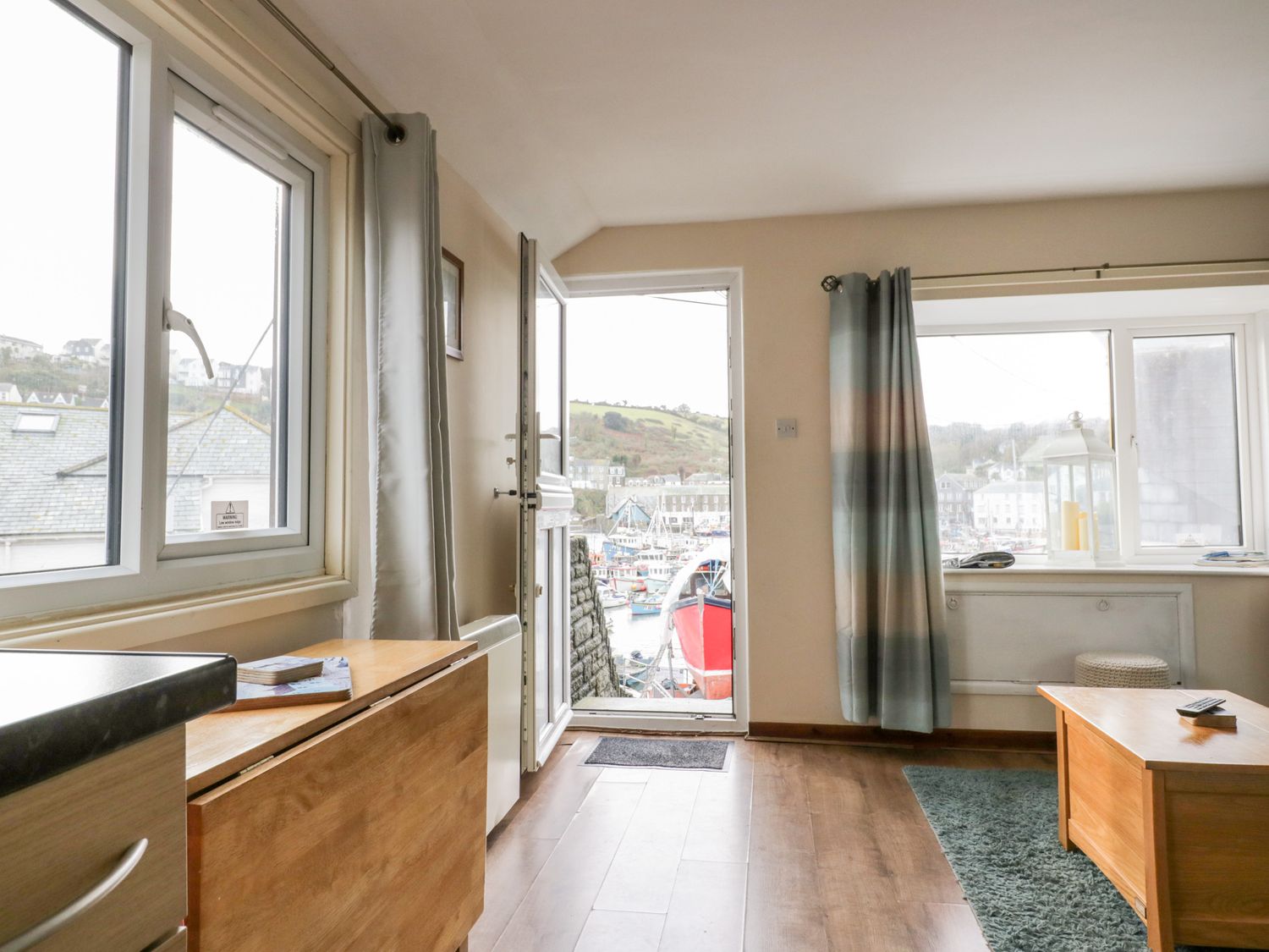 A room with wooden floor a table a door open to a view of boats outside and windows with curtains at The Hideaway in Mevagissey